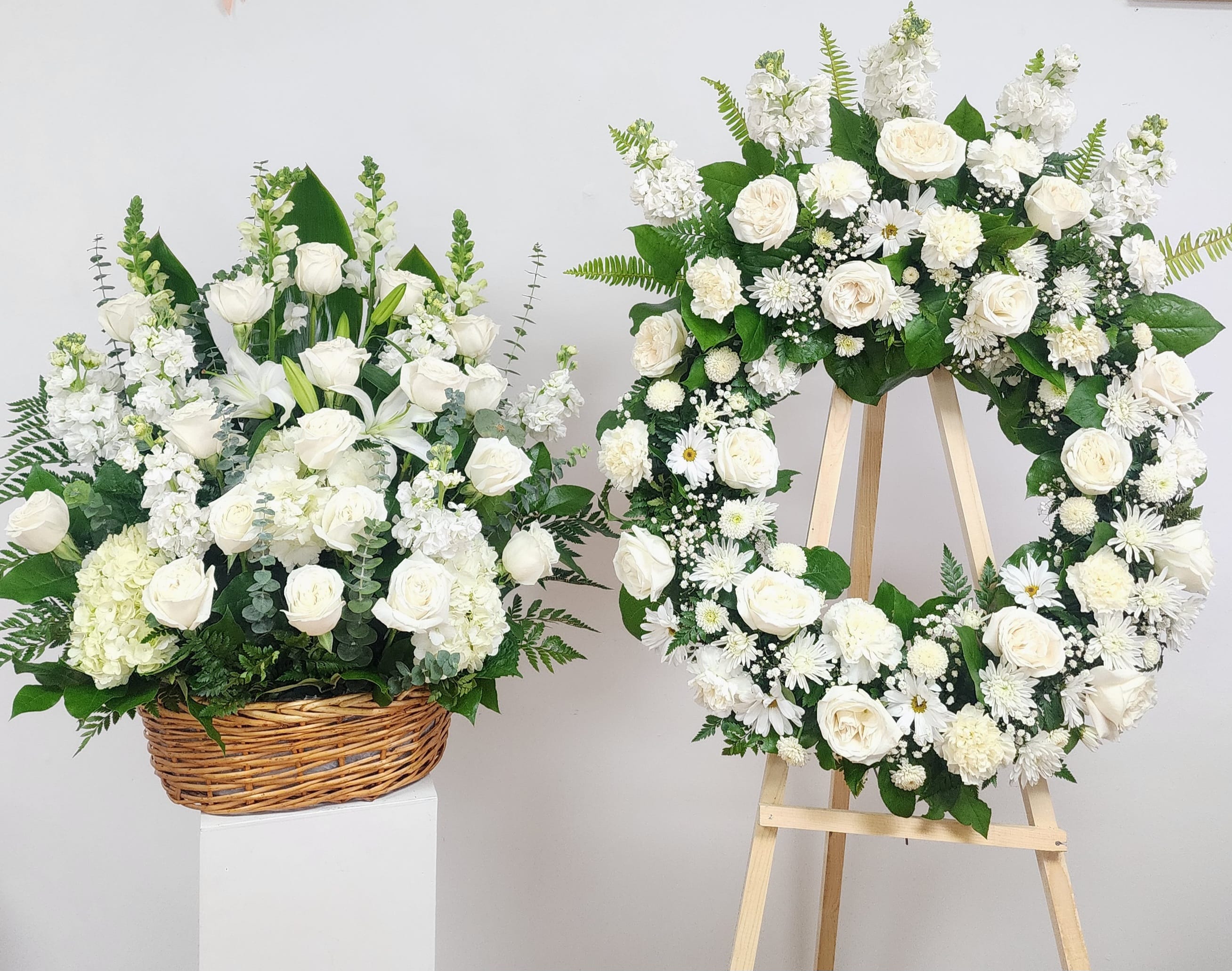White floral wreath on an easel beside a white flower basket arrangement