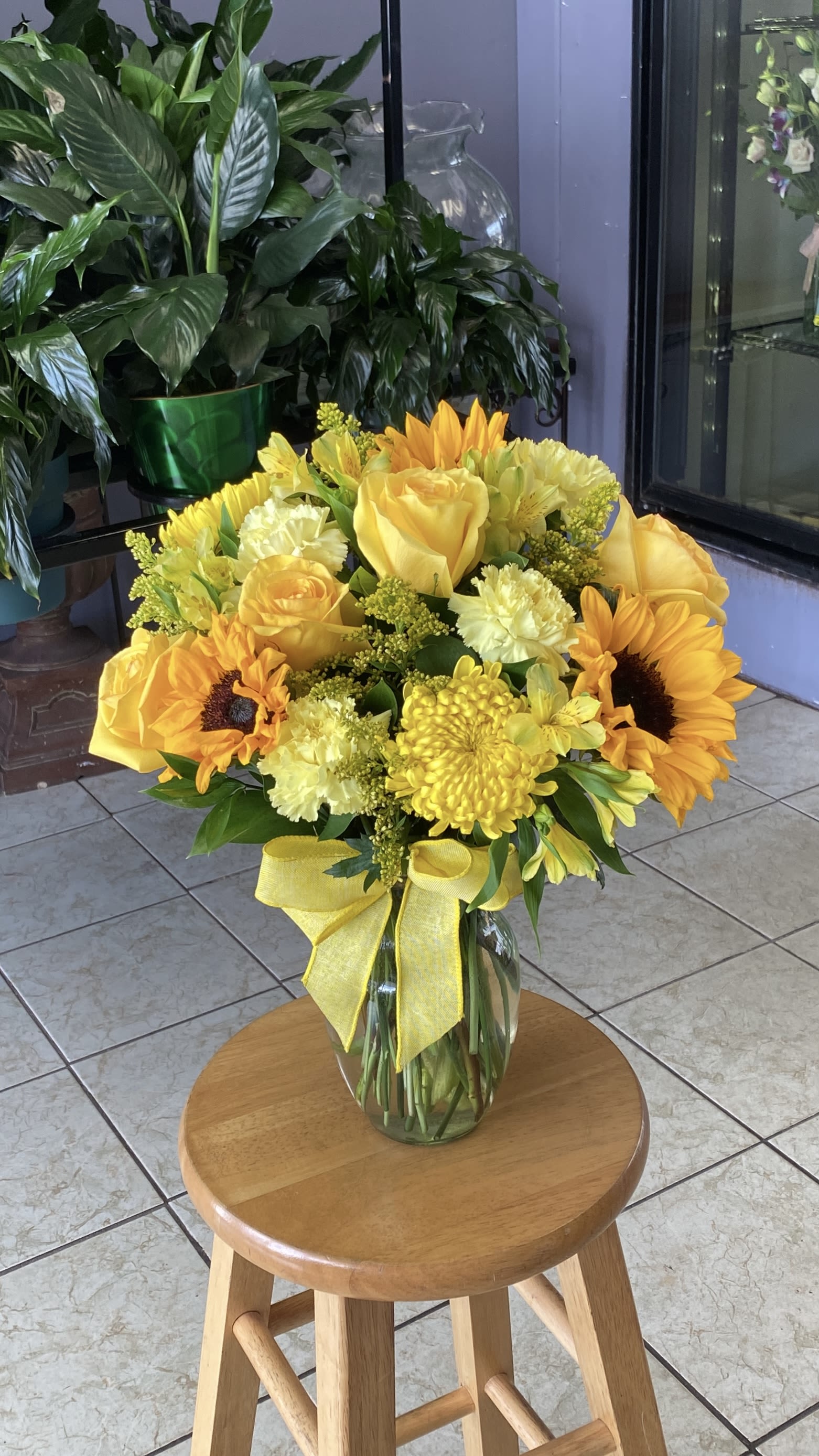 Yellow bouquet of roses, sunflowers, and mums in a glass vase with a ribbon