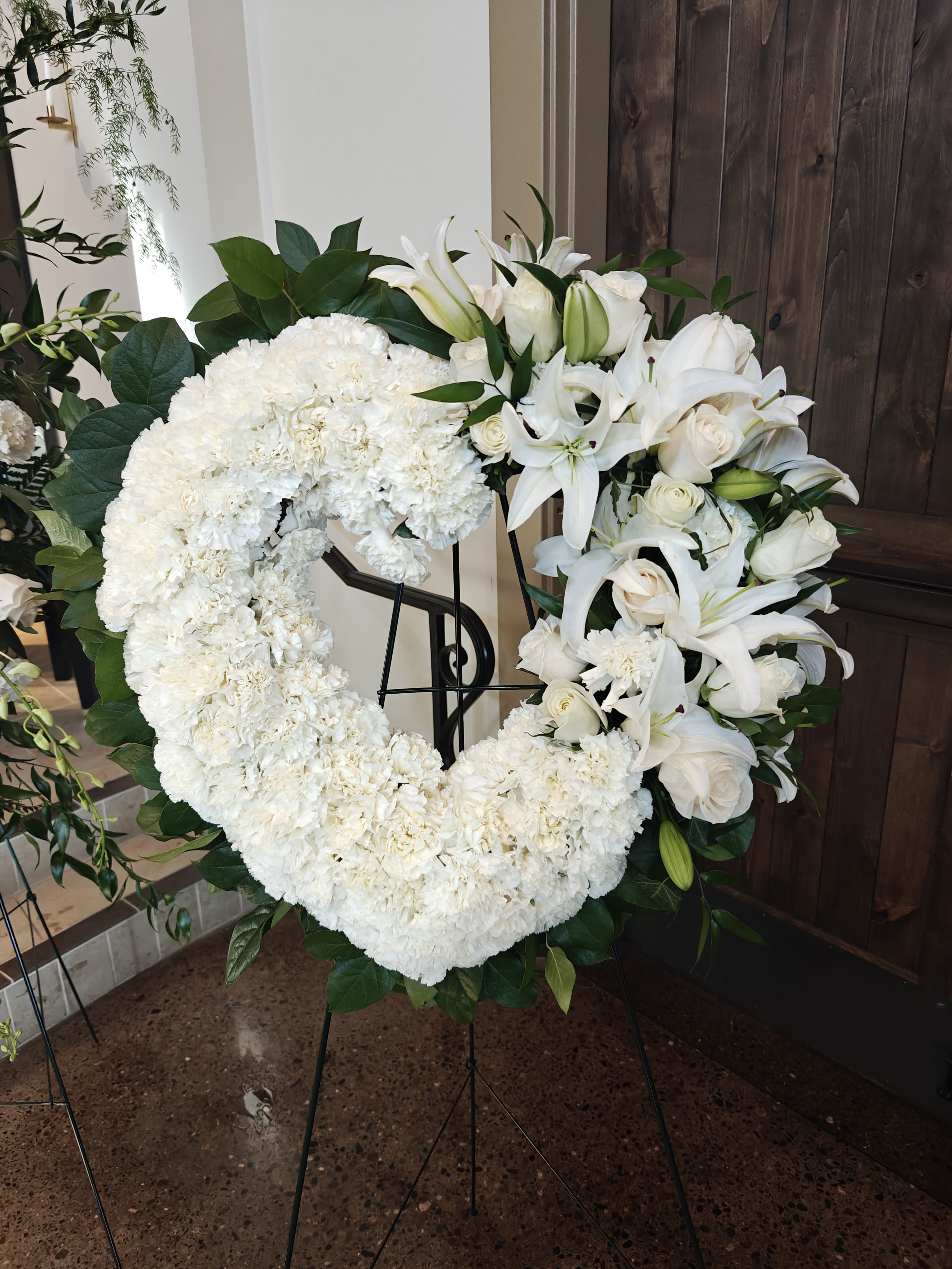 Carnation Heart with Lily Rose Accent - White or Choose another Color - Standard size heart wreath on an easel. We carry larger sizes too. A section of the heart will be dedicated to lovely white lilies and roses.  A classic tribute with some very special flowers. Often this arrangement will be fragrant due to the carnations and Oriental lilies. Sometimes we must substitute the fragrant lilies with standard Asiatic lilies depending on availability. Be sure to order at least a week ahead so that we can bring in plenty of these flowers. It takes almost 100 carnations to make this design.    YES, we can use a different color, at your request, when you order well in advance. 
