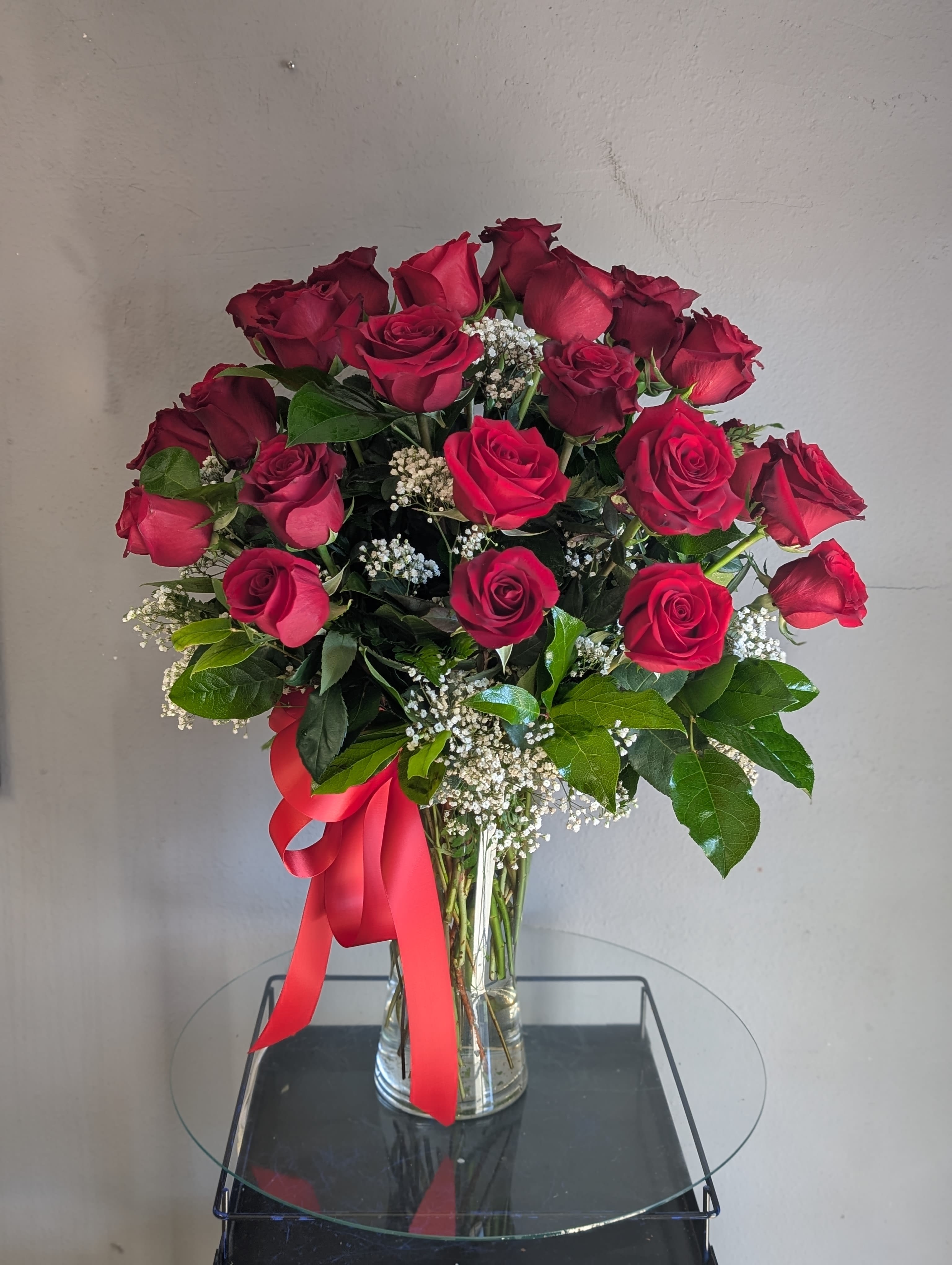 Tall arrangement of red roses with white filler flowers in a clear vase with a red ribbon bow