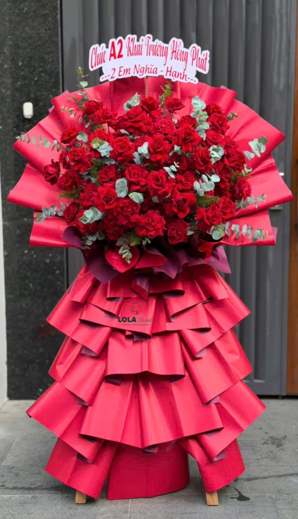 Grand Success Stand - An oversized standing bouquet that reads like a celebration: the Grand Success Stand is a grand congratulatory flower arrangement perfect for business openings, milestone celebrations, or making someone feel honored on a big birthday or anniversary. The dense, domed mass of roses, spray roses, and round chrysanthemums creates a lush focal crown in shades of pink, peach, red, cream, yellow and white, with touches of silvery green foliage giving depth and contrast. The overall effect is joyful and commanding-she'll feel truly celebrated when this arrives.  This design pairs tightly massed blooms with dramatic, tiered wrapping to create a sculptural, dress-like silhouette on a simple wooden stand. Layers of coral and decorative red wrapping paper and soft pink tissue cascade down in ruffled folds, finished with pink and white satin ribbons and long pastel streamers; a printed sign at the top adds a formal congratulatory message. Sized for bold visual impact, it's meant to be the centerpiece at an opening, ceremony, or festive reception-no special care is needed beyond standard flower upkeep, and the wooden stand and structured wrapping conceal the mechanics so the arrangement looks impeccable for the duration of your event.