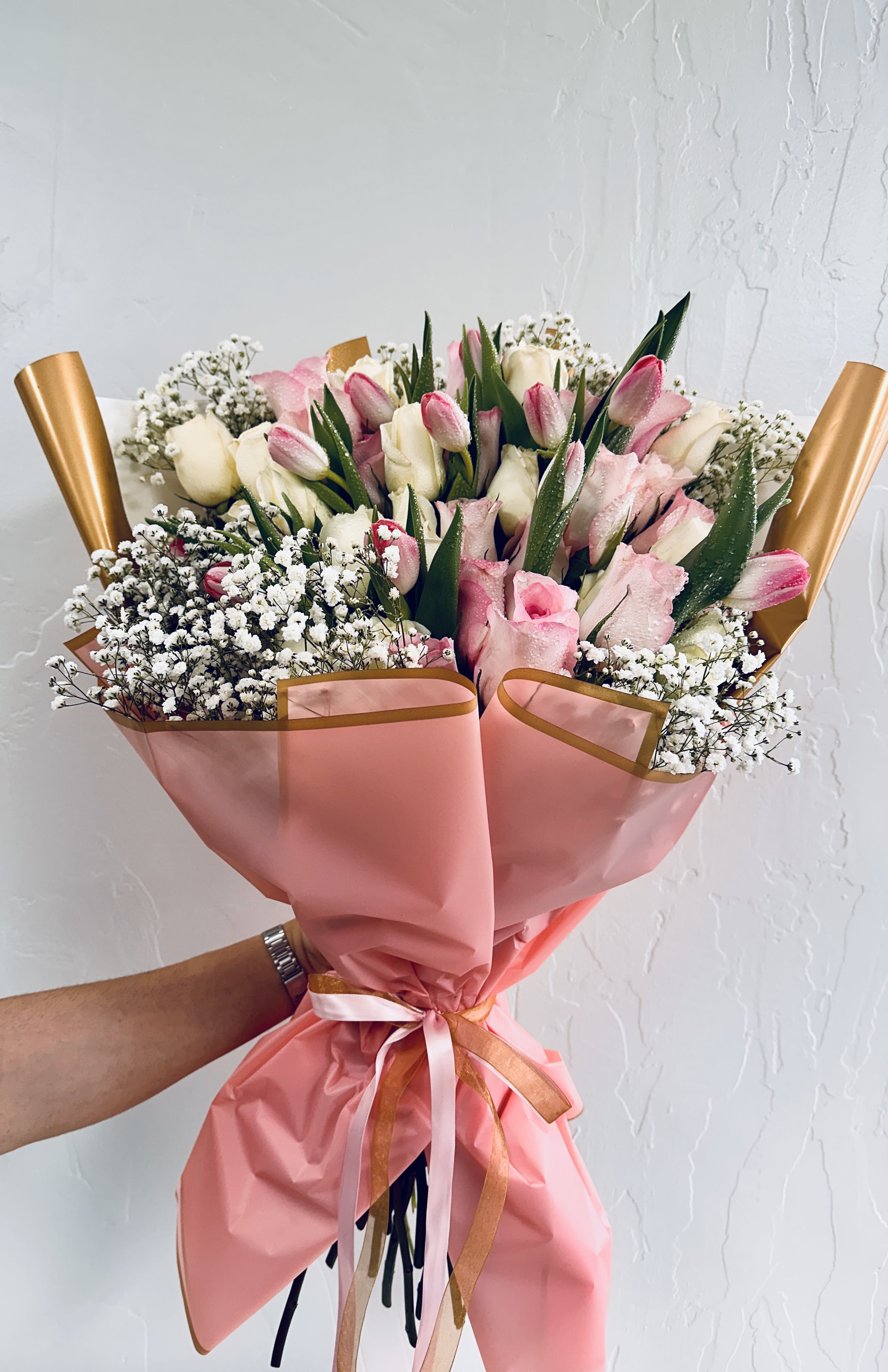 Blush &amp; Whimsy Elegance by The Flower Spot™ - A lush, hand-tied bouquet that reads like a love letter-Blush &amp; Whimsy Elegance by The Flower Spot™ is a pink-and-white flower arrangement perfect for anniversaries, birthdays, graduations, or those "just because" moments when you want someone to feel truly seen. The rounded, full shape and soft palette send a message of romance and celebration, making it an especially lovely choice for love and romance, Mother's Day, Valentine's Day, or a thoughtful thinking-of-you surprise.  This arrangement pairs dense clusters of a dozen pink roses, a dozen white roses and a dozen white or pink tulips with delicate white filler flowers for a soft, airy texture and richly layered visual effect. Finished in soft pink and gold trimming-pink wrapping paper with gold edging, decorative gold wrapping paper, clear plastic protective wrap and a coordinating ribbon-the presentation is polished and gift-ready. The bouquet reads medium to large, with a lush, rounded hand-tied composition that feels curated and personal, as if each stem were chosen and placed by the designer.  Give this bouquet to the person who deserves a moment of delight: a graduate stepping into a new chapter, a best friend for Best Friends Day, or someone you want to celebrate with a romantic gesture. Care is simple-keep stems in fresh water and trim a little off the ends every few days to extend vase life-so the recipient can enjoy its gentle scent and lasting beauty for as long as possible.
