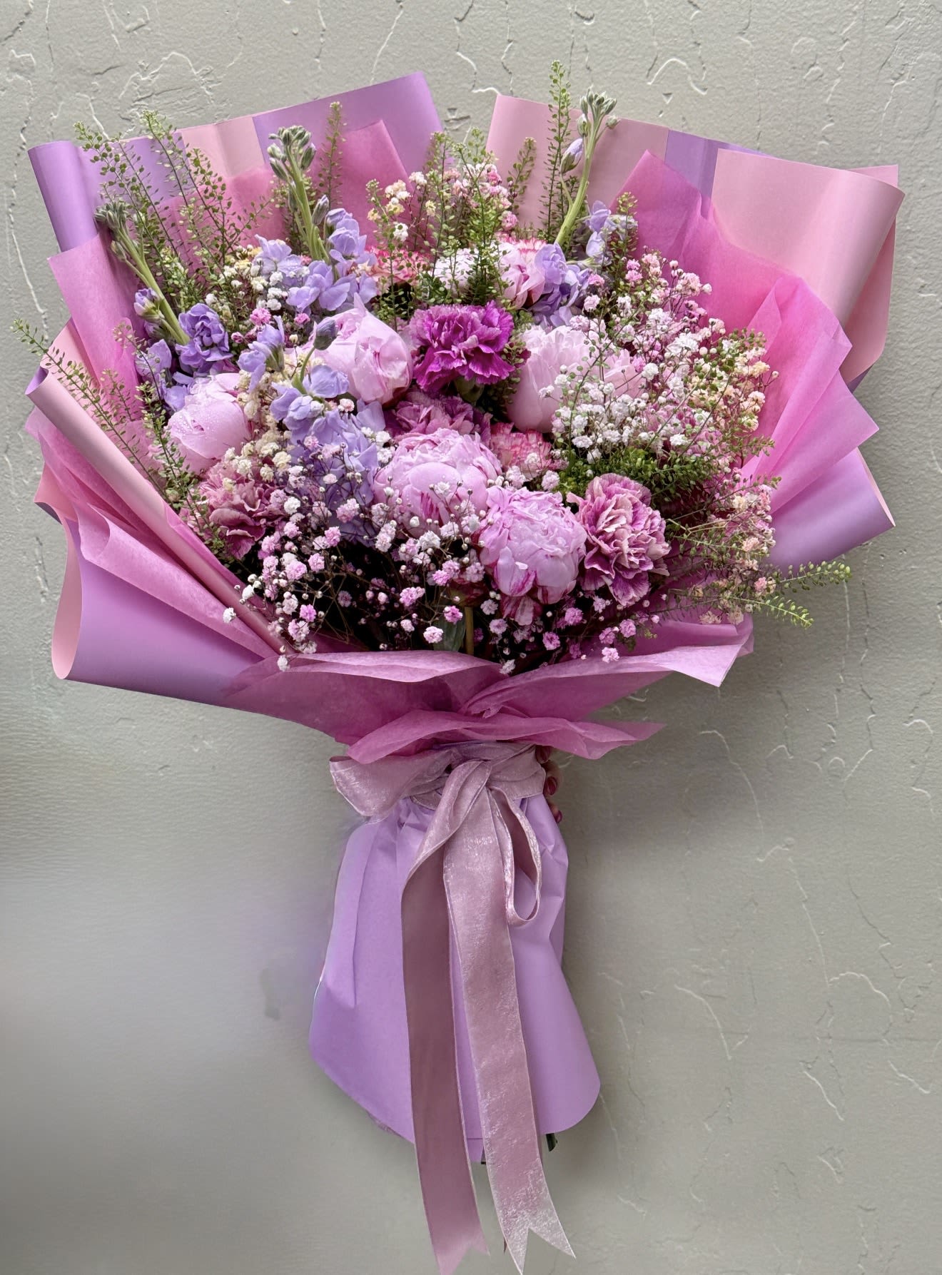 Aura Dream by The Flower Spot - Indulge in the ethereal beauty of Aura Dream, an avant-garde floral masterpiece designed for the discerning aesthetic.This curated arrangement balances rich magenta carnations with rare, cloud-soft blush peonies. Delicate sprays of baby's breath and architectural greenery elevate the texture. Each stem is meticulously selected for its premium quality and vibrant depth. The bouquet is presented in sculptural layers of signature lavender wrapping and anchored by a heavyweight satin ribbon. Aura Dream delivers an unparalleled sensory experience. It is an exquisite statement of modern luxury, tailored for high-profile celebrations, gallery-worthy spaces, or an unforgettable gesture of refinement.