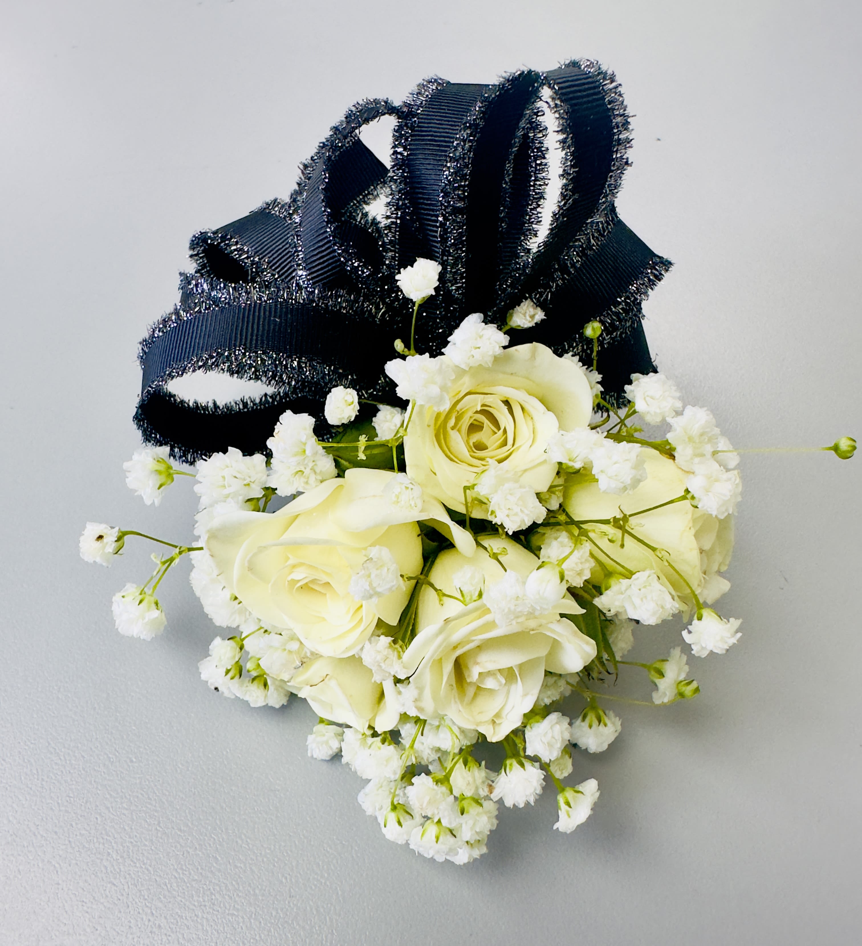 White Rose Corsage by Almaden Valley Flowers™  - Creamy white roses clustered into a compact, hand-tied corsage give this wrist piece a quietly luxurious feel-perfect as a prom accessory or a wedding-day accent that makes the person wearing it feel beautifully noticed. The pale, refined palette reads romantic and timeless, bringing a touch of formal elegance to bridal parties, prom dates, or anniversary looks without overwhelming the outfit.  Each rose is paired with delicate white filler blooms and variegated green-and-yellow foliage for soft contrast and a polished silhouette. Some variations introduce tiny pink accents or airy baby's breath against a dramatic dark ribbon or woven mesh backdrop, and the design is finished with a sheer white ribbon and decorative mesh and bows for comfortable, refined wear. The florist-curated handwork gives each corsage a personal, handcrafted touch.  This is the kind of corsage that suits ceremonies, formal photos, or intimate celebrations-and it remains versatile for sympathy or thank-you gestures where understated grace is desired. Order for same-day or event delivery when available; store cool and mist lightly until wear to keep blooms fresh.