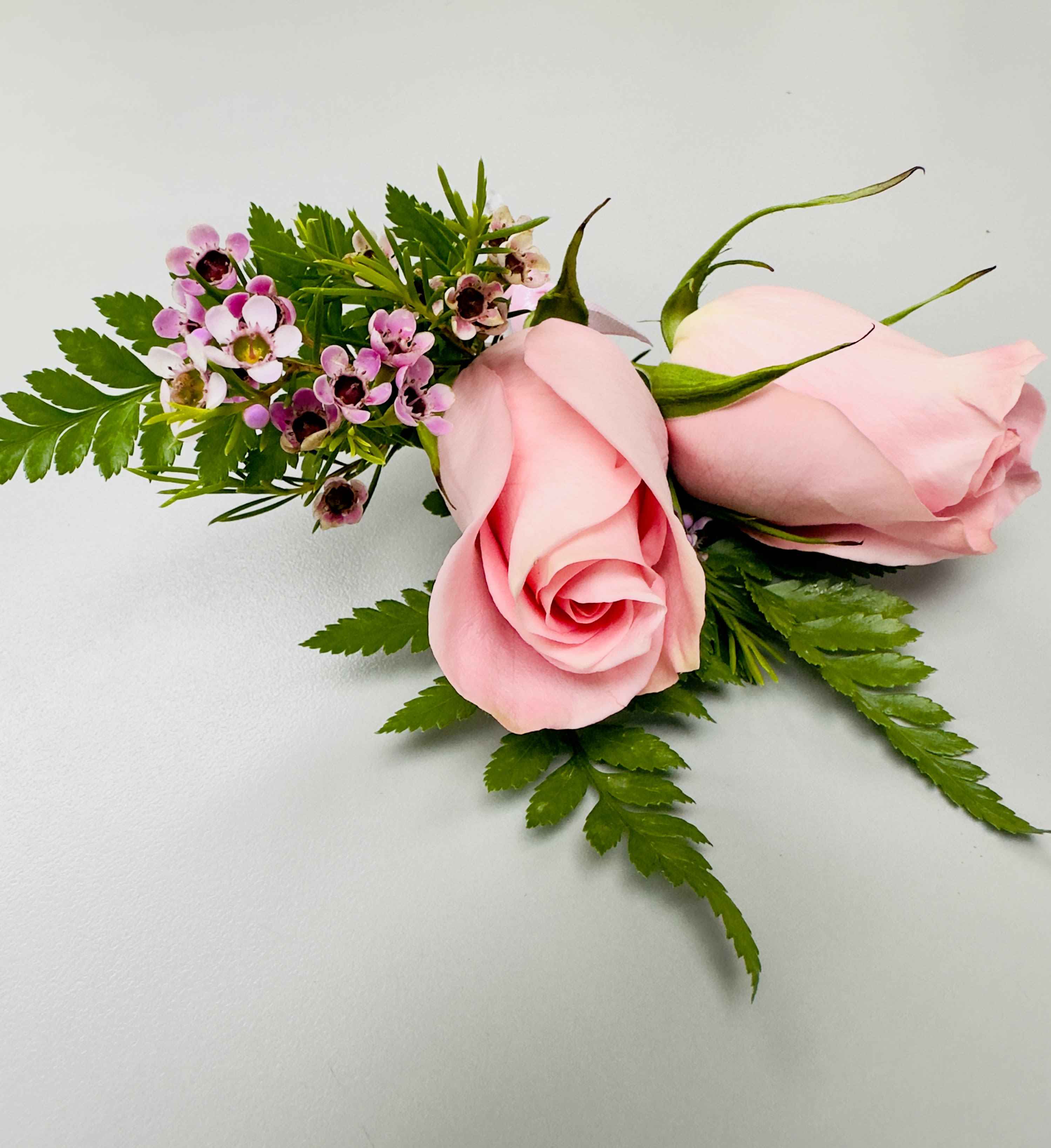 Pink Rose Boutonnière by Almaden Valley Flowers™  - A soft pink rose and a matching rosebud form the romantic focal point of this boutonnière, accented with tiny purple and white filler blooms for a refined, feminine look-perfect for a wedding boutonniere or prom accessory. The compact, wearable scale and delicate pastel palette make the recipient feel thoughtfully styled without overwhelming their outfit.  Long, pointed green leaves and fine textured foliage give the piece a slightly modern, spiky silhouette that contrasts the roses' velvet softness. Wrapped with pale pink satin ribbon and finished with a floral pin, each boutonnière is hand-curated by our designer for a personal, luxury touch that complements bridal parties, groomsmen, or anyone dressing up for a special dance.  Ideal for couples, attendants, or that special student heading to prom, this boutonnière reads as tender and elegant-subtle enough for everyday gifting but polished for formal occasions. Store chilled until wear and use the included pin to secure it to a lapel for best results.