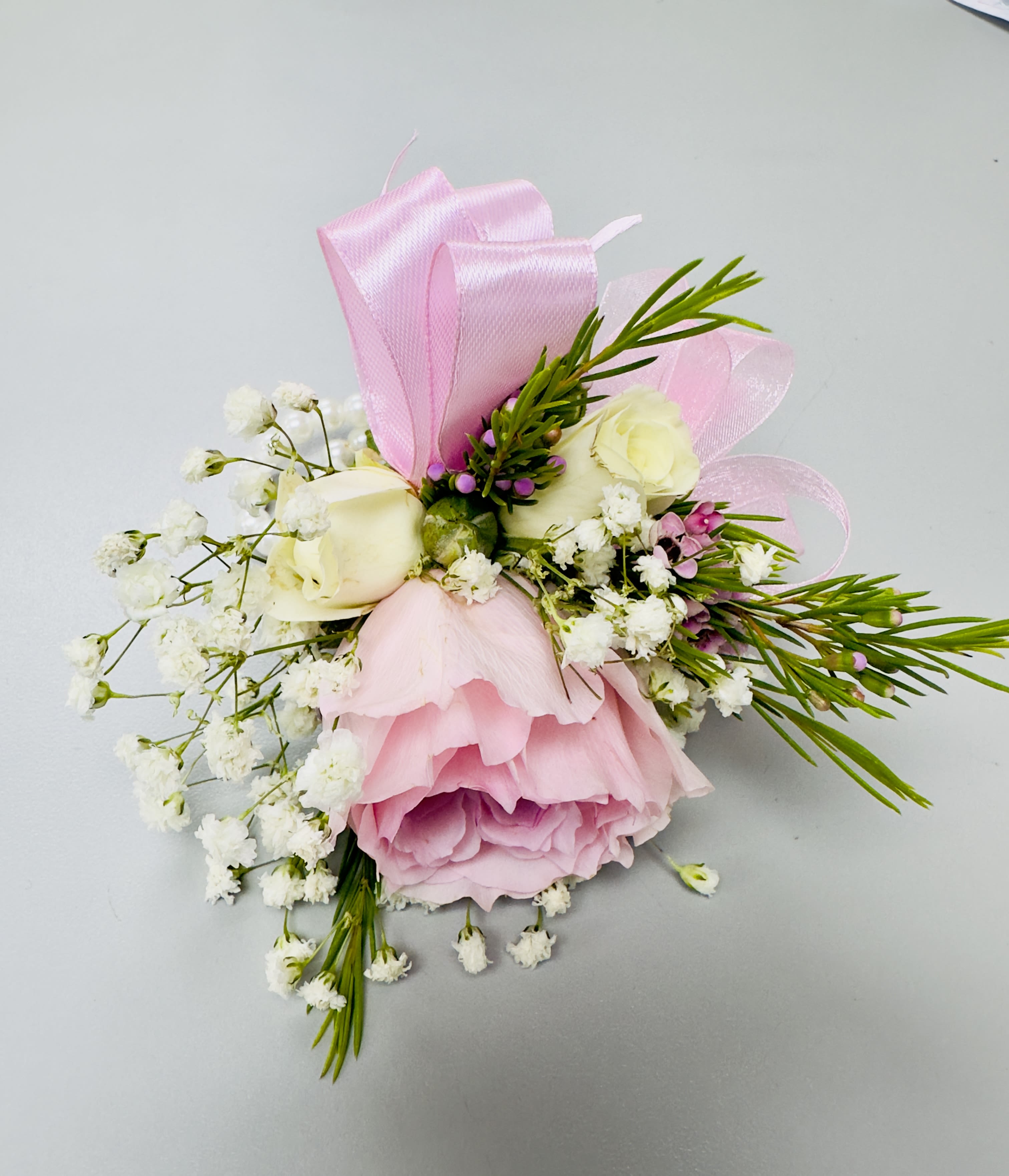 Pink and White Rose Corsage by Almaden Valley Flowers™  - Blush-pink roses clustered with pale spray roses form the heart of this compact corsage, a soft and romantic floral accessory perfect for prom or a wedding. The tight grouping of petals, tiny purple filler blooms and ivory accents create a delicate, feminine look that will make the person wearing it feel noticed and celebrated rather than simply accessorized.  Thoughtfully curated by our designer, each bloom and stem is placed for balance and secured with layered pale pink satin and sheer ribbons that loop through the piece for a refined finish. Glossy green leaves, slender green sprigs and light white filler (baby's breath-style) add airy texture and contrast to the pink and ivory palette; sized to sit comfortably on the wrist or lapel, this wearable arrangement reads elegant and traditional rather than bulky.  Ideal for bridal parties, prom dates, or any formal occasion where you want the recipient to feel truly seen, this corsage arrives ready to wear. Handle gently, keep cool, and avoid direct sunlight to preserve freshness until the moment it's worn.