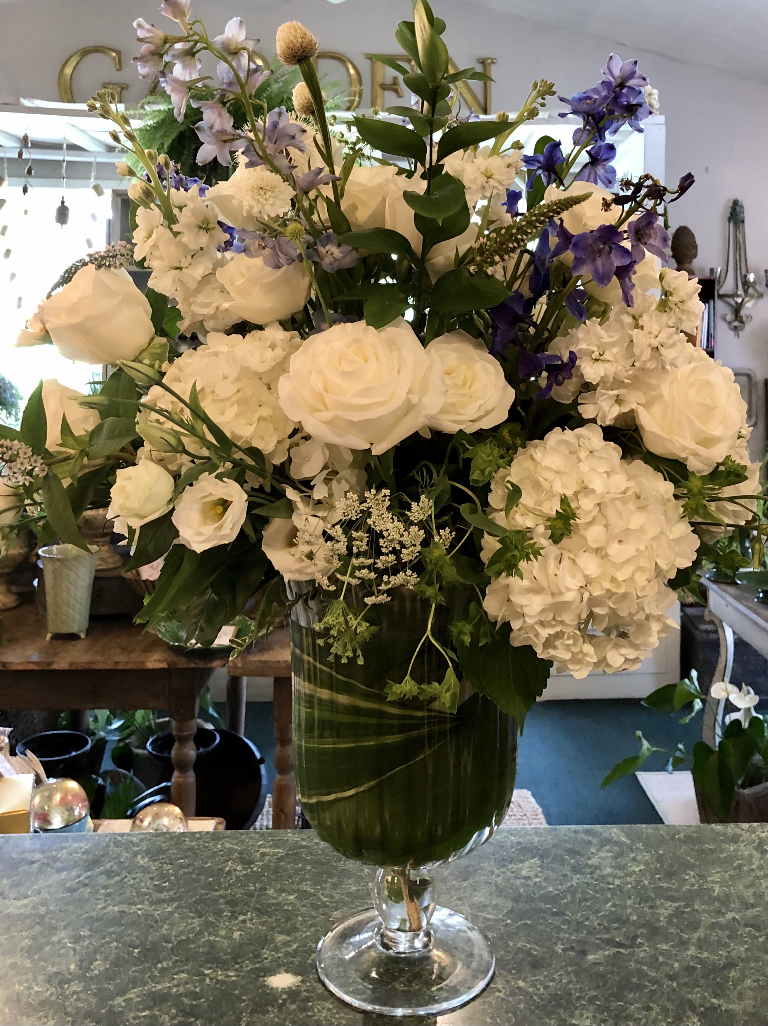Tall arrangement of white roses and hydrangeas with purple delphinium in a clear glass pedestal vase