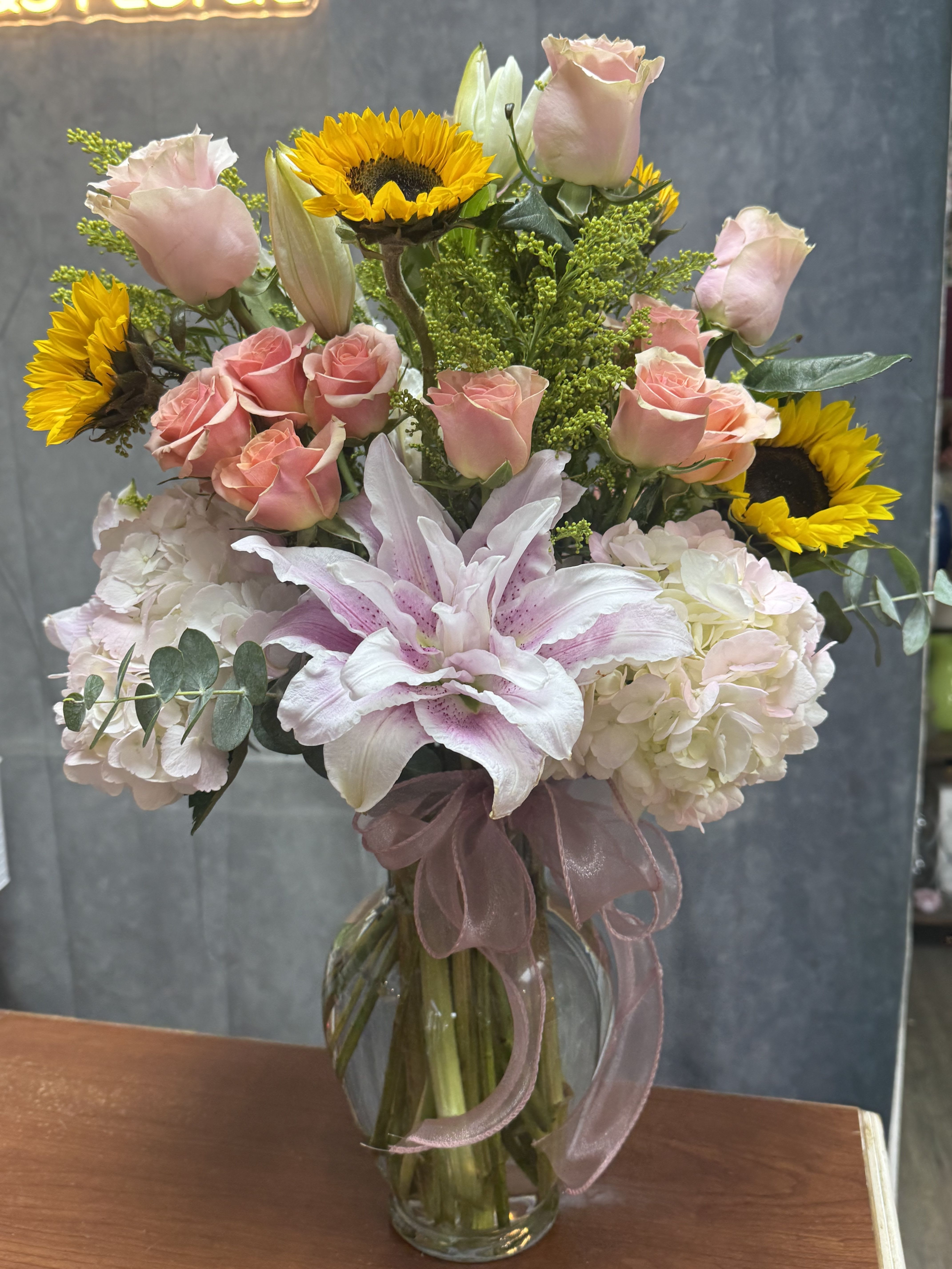 Tall mixed bouquet of pink roses, lilies, hydrangeas and yellow sunflowers in a clear glass vase with ribbon