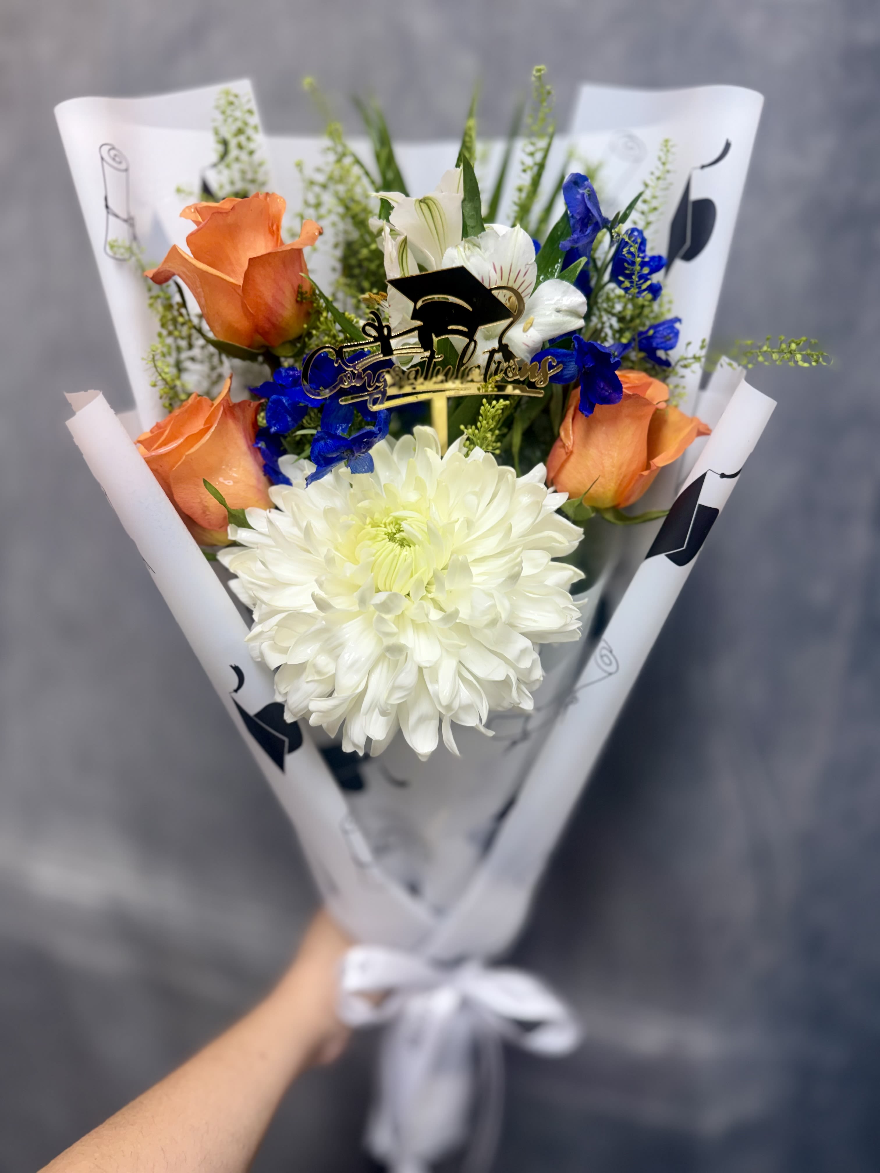 Cap &amp; Bloom bouquet - Bright orange roses with a large white chrysanthemum and vivid blue accents, hand-wrapped in printed white paper with a satin ribbon-cheerful, polished gift for graduations, birthdays, or thanks.