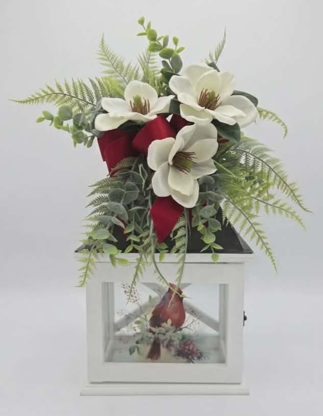 Red Cardinal White Wood Lighted Lantern  - Crisp white magnolia-style silk blooms crown a classic white lantern in this Red Cardinal White Wood Lighted Lantern arrangement, making it an elegant choice for sympathy tributes or thoughtful gifting. The arrangement reads as a refined display the lantern's soft glow paired with deep red ribbon accents creates a quiet, comforting presence perfect for expressing sympathy or offering solace.  Airy silk fern and eucalyptus-like greenery cascade around the lantern, giving the design a gentle, flowing silhouette and soft green contrast to the bright white petals. Rich red fabric woven through the arrangement provides striking color contrast and a formal, polished finish. The included white wood lantern serves as both a decorative accent and keepsake, making this more than just cut flowers. Comes with a battery operated candle ( batteries included).   This is the kind of gift that helps the recipient feel seen and comforted - appropriate for a memorial, a bedside touch, or a seasonal centerpiece that brings warmth to any room. The arrangement arrives ready to display. 13" Tall 9" Wide 