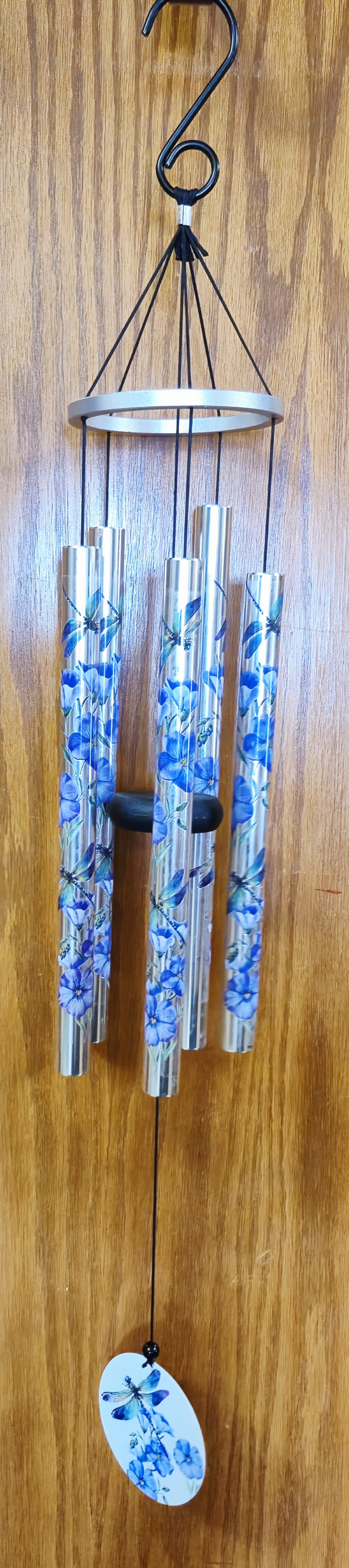 Dragonfly with Purple Flowers Wind Chime - A luminous dragonfly motif and cool blue-and-purple palette make this decorative wind chime a charming gift for birthdays, housewarmings, or as a thoughtful sympathy accent. As a hanging décor piece rather than a traditional flower arrangement, the round white plaque printed with an iridescent blue, purple, and green-winged dragonfly and stylized blossoms immediately catches the eye and sets a serene, whimsical tone for the home or garden.  The piece features silver metal chime tubes printed with coordinating blue floral and butterfly patterns, suspended from a black cord and metal ring. A small oval pendant repeating the botanical motif hangs at the bottom, and the set includes a black hanging hook for easy installation. Measuring 36" from hook to paddle, the design balances visual detail with airy movement and a cool lavender and white color story that complements indoor or covered outdoor spaces.  Perfect for the person who appreciates calming, artistic accents, this wind chime sends a message of gentle comfort and thoughtful remembrance without being overt. It arrives ready to hang - choose a sheltered porch, entryway, or memorial site where soft breezes can let the chimes sing.  Choose the Deluxe presentation option to include the stand with your purchase for presentation use at a service or visitation.