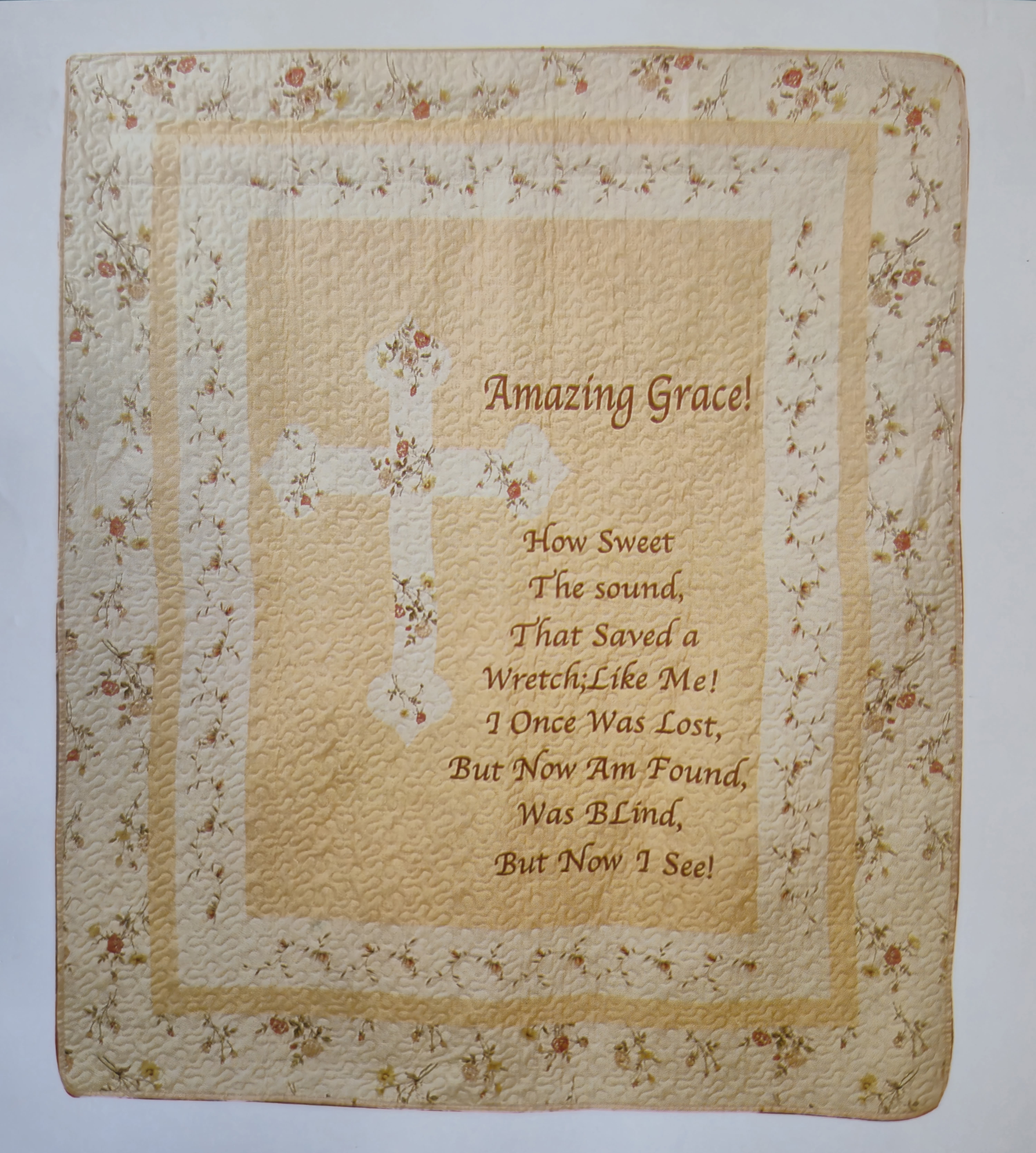 Amazing Grace Cream Throw  - A soft cream quilted throw embroidered with a printed cross and the words "Amazing Grace" makes a comforting sympathy gift or devotional keepsake for someone grieving or remembering a loved one. This devotional throw and wall-hanging-style design suits a memorial service, bedside comfort, or a quiet anniversary of remembrance, offering a gentle visual of faith and reassurance to the person you're thinking of.  Made in warm cream and pale yellow tones with a delicate small-floral border, the 50 x 60-inch throw has a handmade, heirloom texture that reads like a cherished family piece. The center panel features the hymn phrase and cross motif in brown lettering; the soft quilted fabric and muted palette keep the look traditional and calming. A coordinating quilted wall hanging or the included simple metal easel stand can display the piece at a service or in the home, turning it into both a practical blanket and a meaningful display item.  Give this as a tender way to say you remember and care-she'll feel comforted by the devotional message and the cozy, tactile warmth. Machine wash gentle or follow included care instructions for best longevity; choose the easel option if you'd like it presented upright for memorials or gatherings.