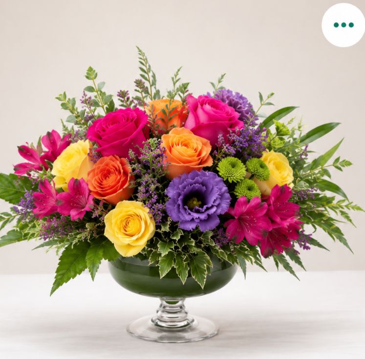 Mosaic  - Bright, energetic color pops make this mixed-color floral arrangement an uplifting choice for an anniversary or any celebration. The arrangement works beautifully as a birthday bouquet, a cheerful get-well surprise, a thinking-of-you gift, or a just-because pick-me-up - its lively palette also suits occasions like graduations, congratulations, and sympathy when you want to offer warmth and comfort.  A full, asymmetrical composition of yellow and red roses, red tulips, purple lisianthus, yellow alstroemeria, and small green chrysanthemum blooms sits in a low glass pedestal bowl. Patterned foliage and blooms extend outward for a dynamic, abundant look that reads joyful rather than formal. The mix of textures - velvety roses, crisp tulip petals, delicate lisianthus, and daisy-like chrysanthemums - creates visual movement and a bright, inviting scent profile.  This is the kind of arrangement that makes the recipient feel seen and celebrated: perfect for saying "happy birthday," "congratulations," "I'm thinking of you," or "I'm sorry." Sized to sit easily on a table or bedside, the glass pedestal bowl presents the flowers at eye level; for best longevity, keep in fresh water, away from direct heat, and recut stems if moving the arrangement to a new vase.