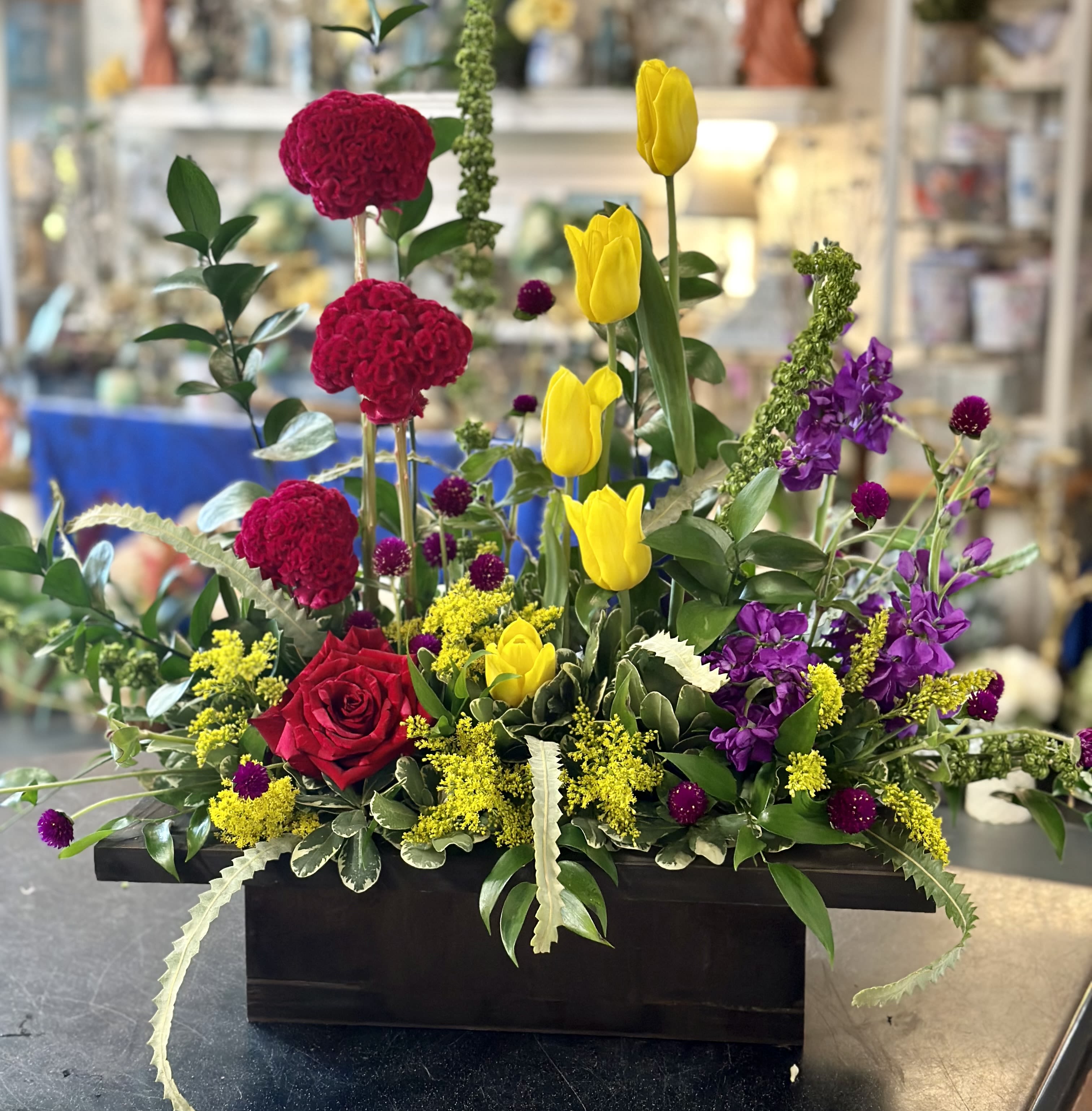 Mixed floral arrangement with red roses, yellow tulips, and purple blooms in a black container