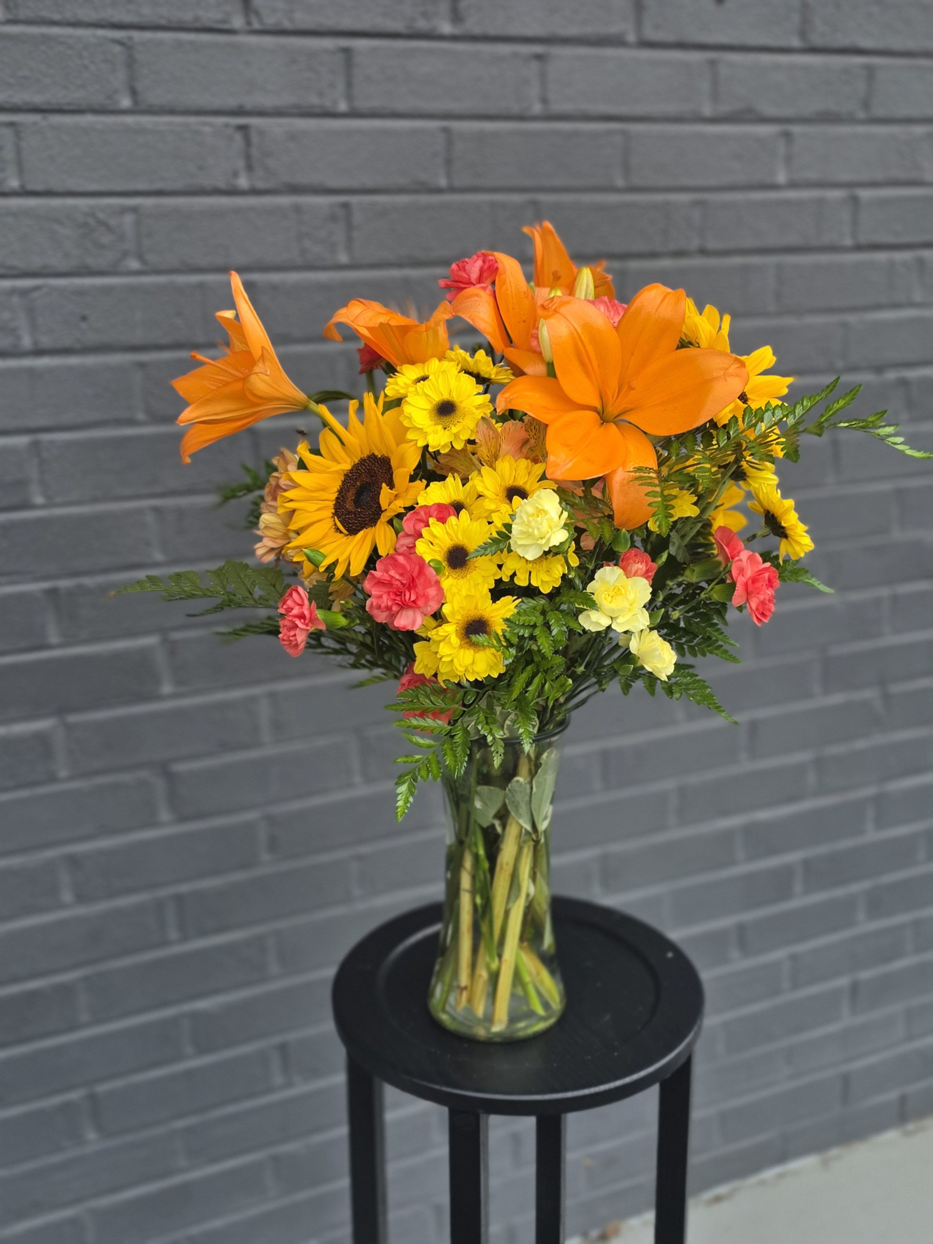Happy Place  - Bright orange lilies take center stage in this cheerful mixed bouquet, making it a vibrant choice for a birthday bouquet, thank-you gift, or general celebration. The tall, trumpet-shaped lilies rise above a garden-style cluster of yellow daisy-like blooms and sunflower faces, while soft pink and pale yellow carnations add gentle contrast so the recipient feels genuinely celebrated by the warmth and energy of the arrangement.  Arranged in a clear glass vase, the stems show through the water for a fresh, casual look that suits everyday gifting as well as springtime events and congratulations. The composition is full and upright, medium-to-large in scale, and features a sunny palette of orange, yellow, pink, green, and white for a lively, summery impression that brightens any room.  This bouquet reads as lively and informal-the kind of arrangement that says "you're thought of" without fuss. For best enjoyment, refresh the water every two days and trim stems slightly when you do; the clear vase and sturdy stems make it easy to keep the display looking just-cut for several days.