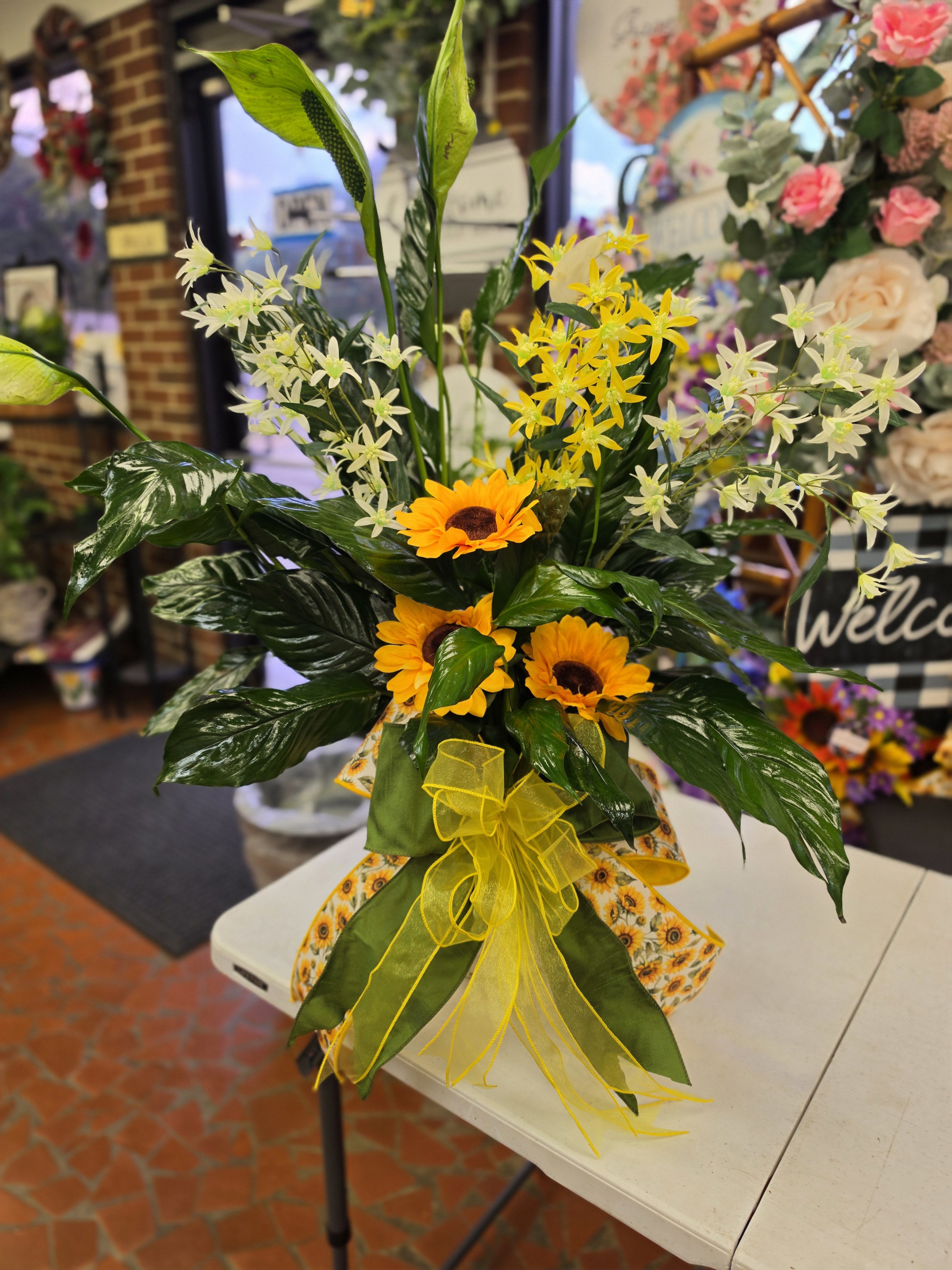 Peace Lily Dressed Up Bright - Three bright sunflower blooms take center stage in this cheerful sunflower bouquet for everyday gifting or a celebratory occasion. The sunny yellow and warm orange tones are balanced by sprays of small white and yellow star-shaped flowers and large glossy green leaves, so the person you're thinking of will feel genuinely uplifted the moment they see it.  Arranged in a wrapped container with patterned decorative paper and finished with a sheer yellow ribbon, this upright, full silhouette reads casual but thoughtfully styled. The tall foliage adds height and texture while the clustered sunflowers provide bold focal points; perfect for birthdays, congratulations, or a just-because surprise. Comes ready to display in its decorative wrap (vase option available) - minimal care needed: fresh water and a cool spot will keep the blooms bright for days.