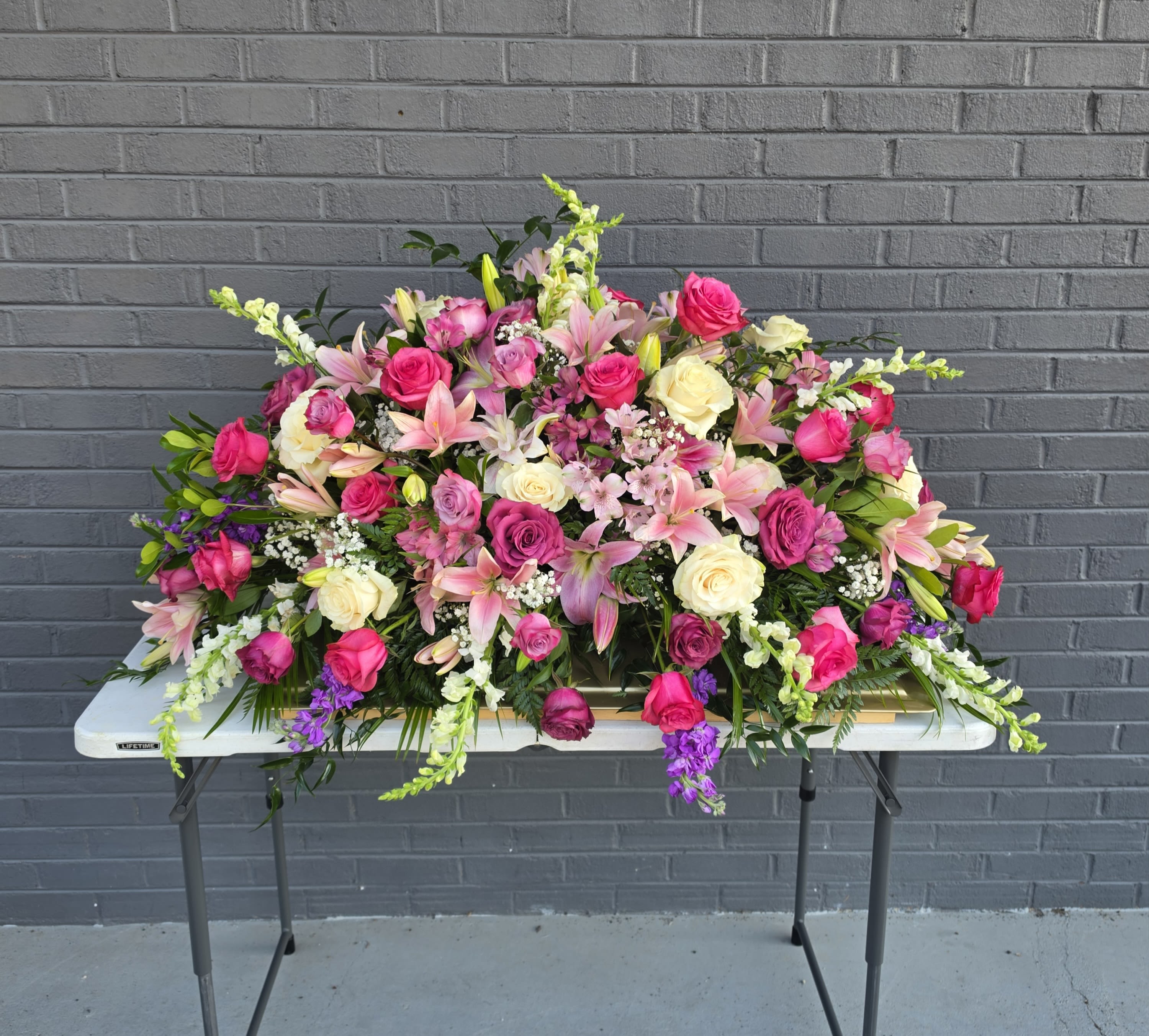 Casket spray Pink Love - A sweeping casket spray designed for sympathy and memorial services, this large funeral arrangement fills the space with abundant pinks and soft creams to convey warmth and remembrance. Rich hot-pink and blush roses paired with pink lilies create a romantic, compassionate impression the person you're honoring will be remembered by, while white blooms and lavender accents lend calm contrast and dignity.  Built low and broad to sit gracefully across a casket or display table, the spray features layers of roses, lilies, tall snapdragon spikes, and delicate white filler flowers woven throughout for texture and movement. Darker foliage and trailing edge blooms give the piece a finished, cascading silhouette; overall dimensions are substantial to provide strong visual presence at a service.  Appropriate for funerals, memorials, or a heartfelt tribute, this arrangement is meant to make mourners feel comforted and seen. Care is handled by the florist for delivery and placement on the casket or table; contact us if you need specific sizing or coordination with service details.