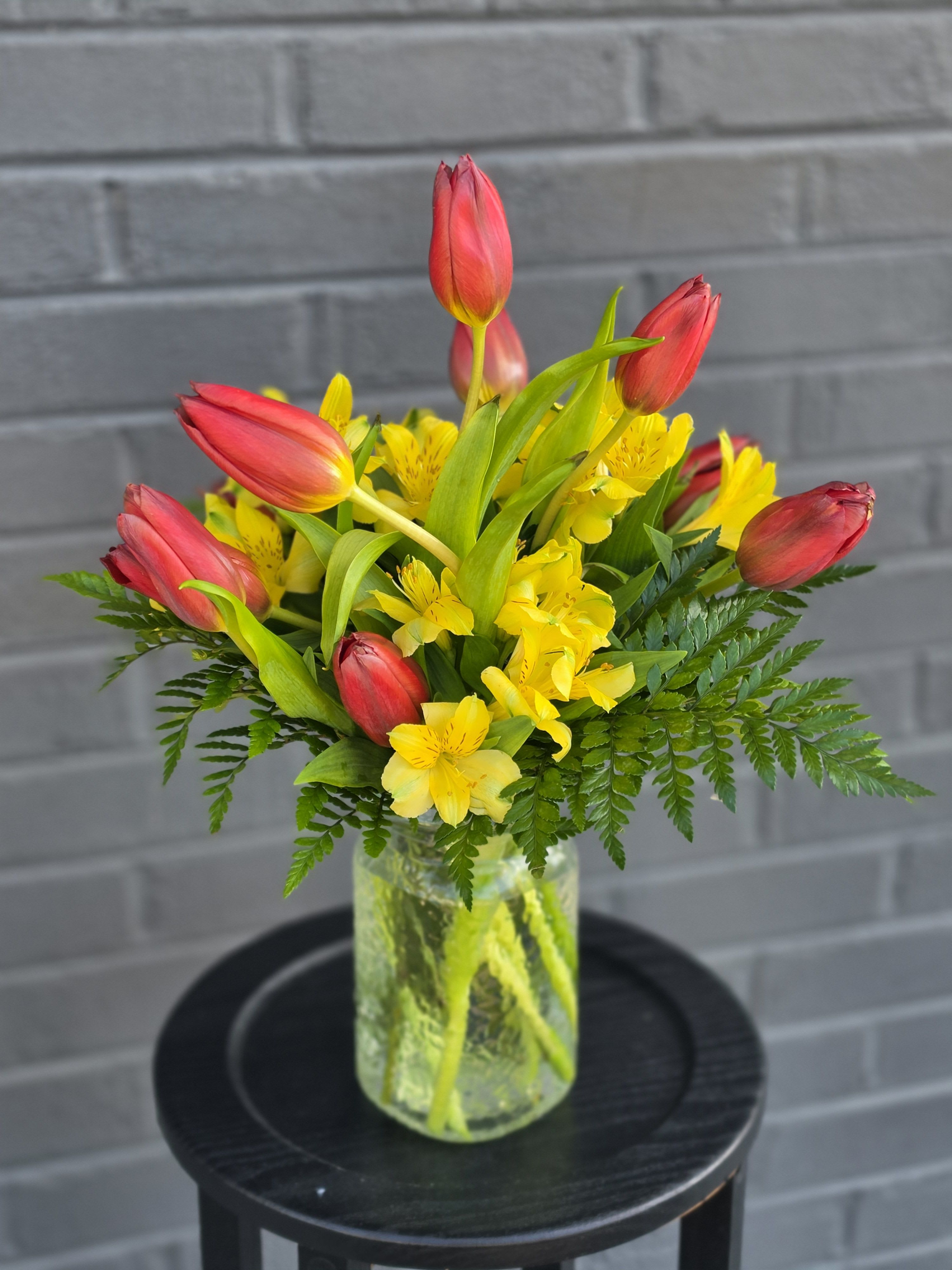 All About You - Upright red tulips paired with sunny yellow alstroemeria create a cheerful spring bouquet perfect for birthdays, thank-you gifts, or just because - a bright everyday arrangement that will make the person you're thinking of feel genuinely celebrated. The bold red-and-yellow contrast reads energetic and hopeful, while the loose, rounded silhouette keeps the look casual and friendly for seasonal displays or celebratory moments.  The flowers are arranged in a clear glass vase so the tall tulip stems can rise above clusters of alstroemeria, with fern-like foliage fanning around the edges to soften the shape. Colors: red, yellow, green; flowers: tulips and alstroemeria; hard good: clear glass vase. The composition is compact yet lively, roughly upright and easy to place on a table or desk - a simple, fresh statement that delivers big cheer with minimal fuss.