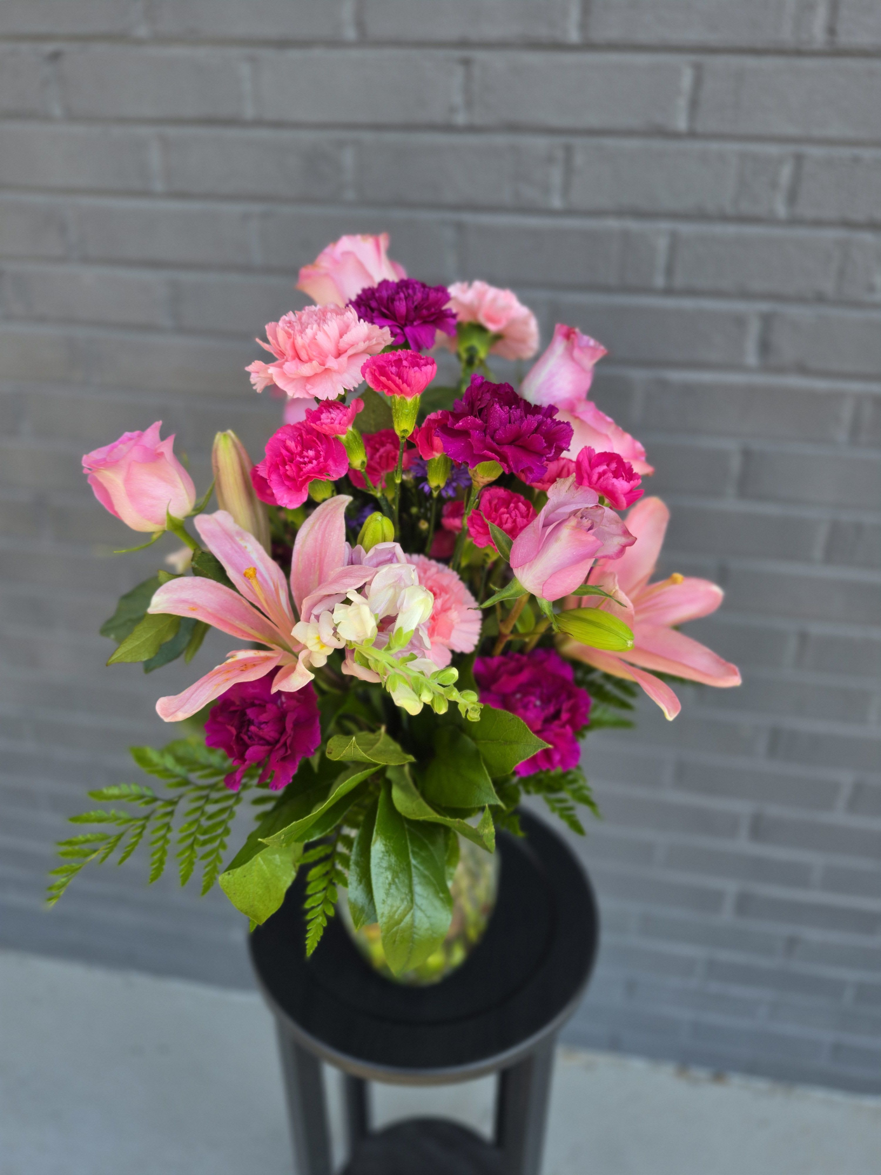 Pink Passion Love - Soft pink lilies, blushing roses, and vivid magenta carnations combine in a cheerful, romantic bouquet perfect for a birthday bouquet, thank-you gift, or springtime celebration. The mix of soft blush and saturated fuchsia creates an immediately uplifting impression-the kind of arrangement that makes the person you're thinking of feel truly seen and celebrated.  This compact, upright design is arranged in a clear glass vase for a simple, fresh presentation. Visible blooms include lilies, rose buds, carnations, snapdragons and small pale white accent flowers, all nestled with fern-like foliage and broad green leaves for a full, rounded shape. The contrast of warm pinks and cool purples adds depth and texture, giving the arrangement a lively, layered look.  Ideal for marking birthdays, saying thanks, or brightening a spring table, this bouquet arrives ready to display. Keep flowers in a cool spot out of direct sun and refresh the water every two days to extend enjoyment - the kind of gift that keeps giving long after the moment it was received.