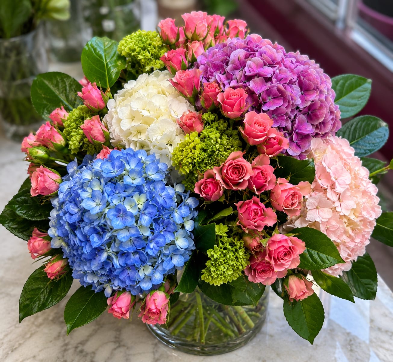 Garden Bliss  - Inspired by the beauty of an English garden in full bloom, Garden Bliss is a lush, handcrafted arrangement designed to make a lasting impression.  This luxurious design showcases stunning blue, lavender, and white hydrangeas paired with delicate blush pink spray roses, vibrant green viburnum, and premium seasonal greenery. The harmonious blend of soft pastels and vivid botanical textures creates a full, elegant arrangement that's both sophisticated and naturally beautiful.  Designed in a premium clear glass vase by the floral artists at Bosmat Flowers, Garden Bliss is perfect for birthdays, anniversaries, housewarmings, thank-you gifts, or simply brightening someone's day. Every arrangement is handcrafted using the freshest blooms available, ensuring exceptional quality and long-lasting beauty.  Whether displayed as a stunning centerpiece or gifted to someone special, Garden Bliss brings freshness, elegance, and timeless charm to any setting.