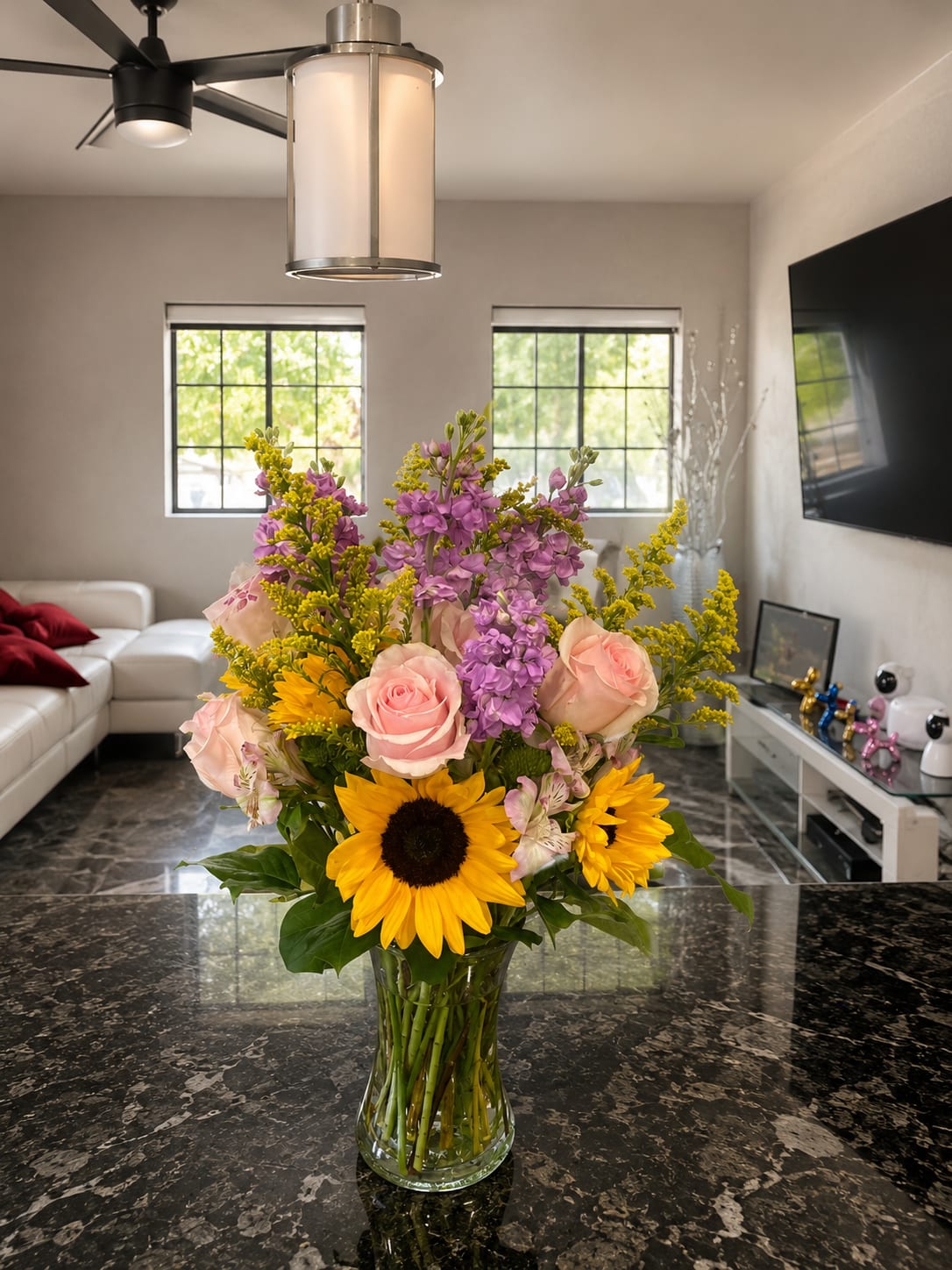 Spring Glow - Bright yellow sunflowers pair with soft pink roses and tall purple filler blooms in a clear glass vase to create a cheerful birthday bouquet or just-because centerpiece. The mix of bold sunflower faces and rounded rose heads gives a joyful, slightly romantic feel-perfect for saying thank you, celebrating a birthday, or surprising someone who needs a sunny pick-me-up.  This medium-to-large arrangement reads as an upright, fresh centerpiece for the home. The design layers airy vertical accents with lush rose clusters and striking sunflower centers for strong color contrast and texture; whites tucked throughout add light and balance. Arrives ready to display in the included clear glass vase-ideal for someone who should feel genuinely celebrated.