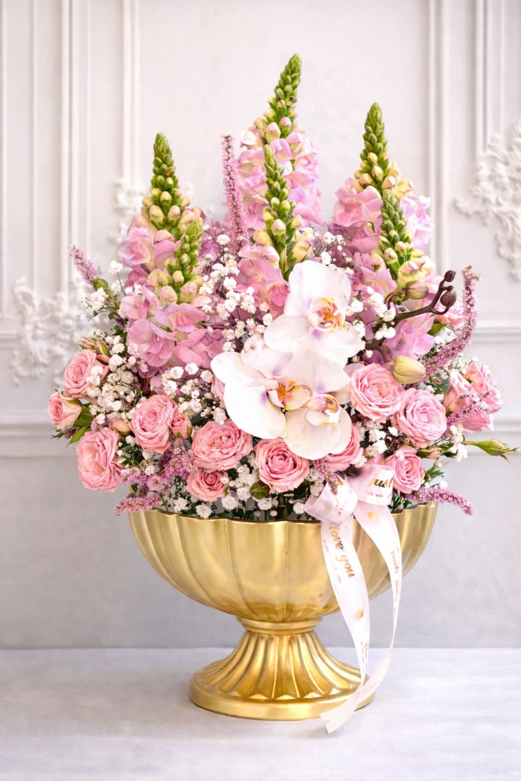 Rosé in Gold -  Rosé in Gold is a luxurious floral statement designed to captivate from every angle. This elegant arrangement features soft pink spray roses, delicate baby’s breath, feathery astilbe, tall snapdragons, and a graceful cascade of white Phalaenopsis orchids, all artfully designed in a reusable gold pedestal vase.  Perfect for Valentine’s Day, anniversaries, grand gestures, and elevated home décor, this arrangement brings romance, height, and timeless beauty into any space. Finished with a soft ribbon detail, Golden Blush Symphony is designed to leave a lasting impression long after the flowers bloom.  Available for delivery in Carlsbad and surrounding North County San Diego areas.