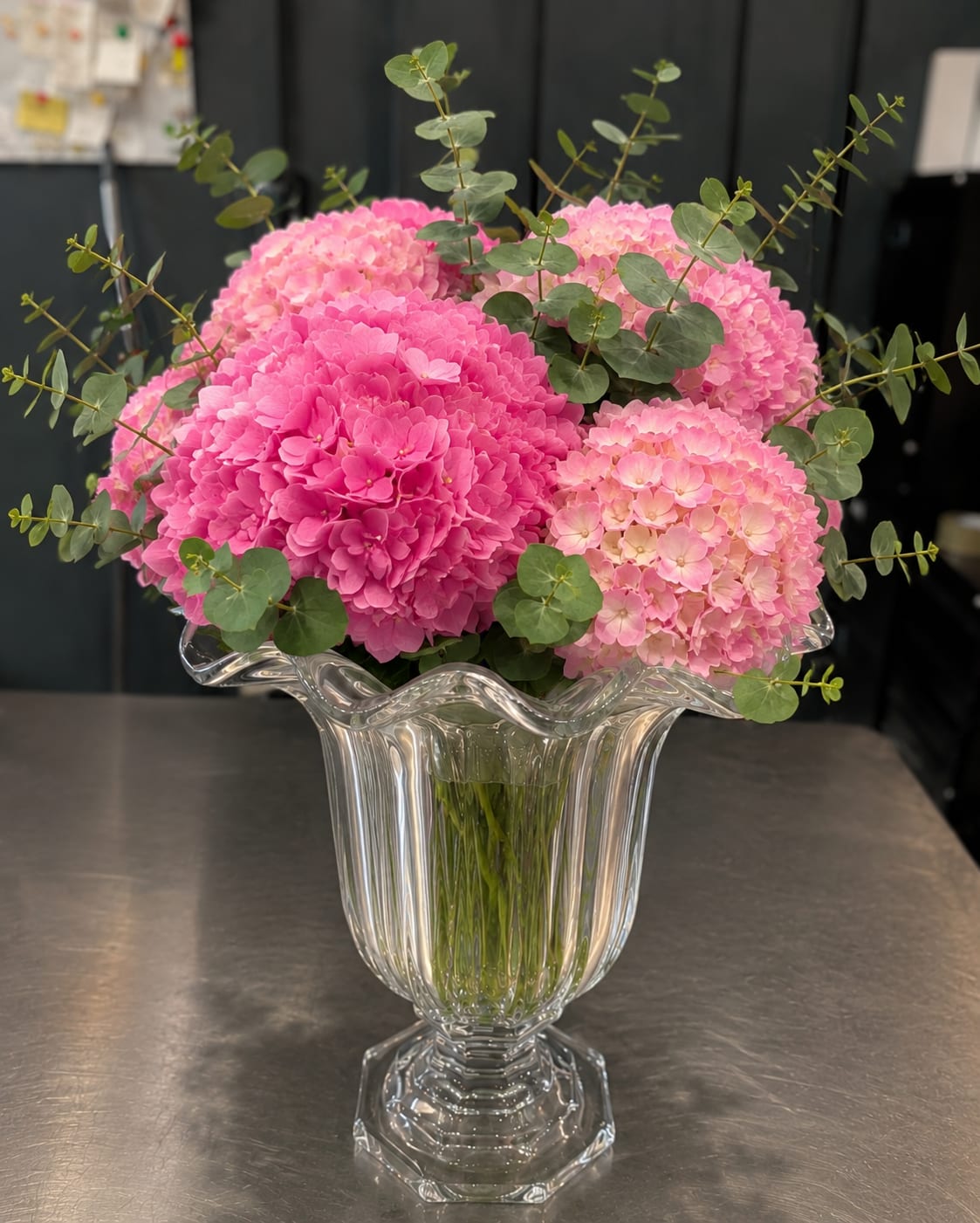 Rose Pearl - Rounded clusters of pink hydrangeas-from bright fuchsia to soft blush-give this flower arrangement an immediate, joyful presence perfect for a birthday bouquet, spring celebration, or just because you're thinking of someone special. The airy eucalyptus stems weave through the dense blooms, adding fresh green contrast and gentle movement so the recipient feels uplifted and truly seen.  Set in a fluted clear glass vase with a pedestal base, the composition reads as both classic and lively: lush white highlights peek between the pinks, while the vase's elegant shape shows off the stems and keeps the look light on a mantel or dining table. Sized to feel abundant without overwhelming a room, it arrives ready to display and makes a cheerful everyday gift that brightens any space with color and texture.