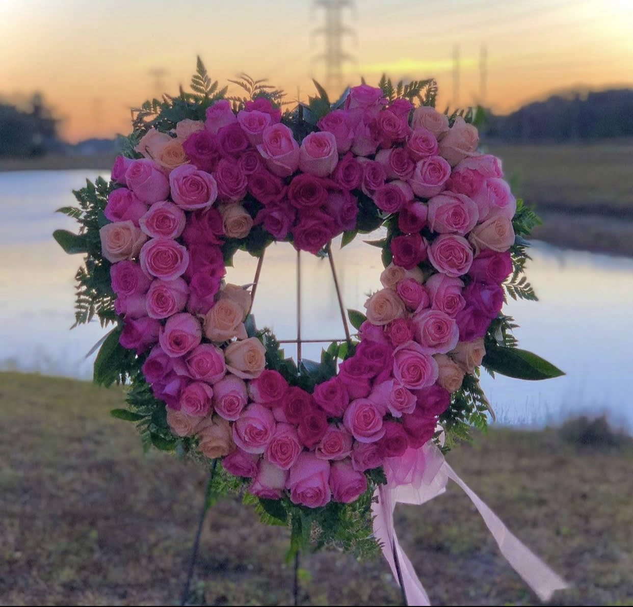 Heart-shaped standing wreath of pink and peach roses on an easel with trailing ribbons outdoors at sunset