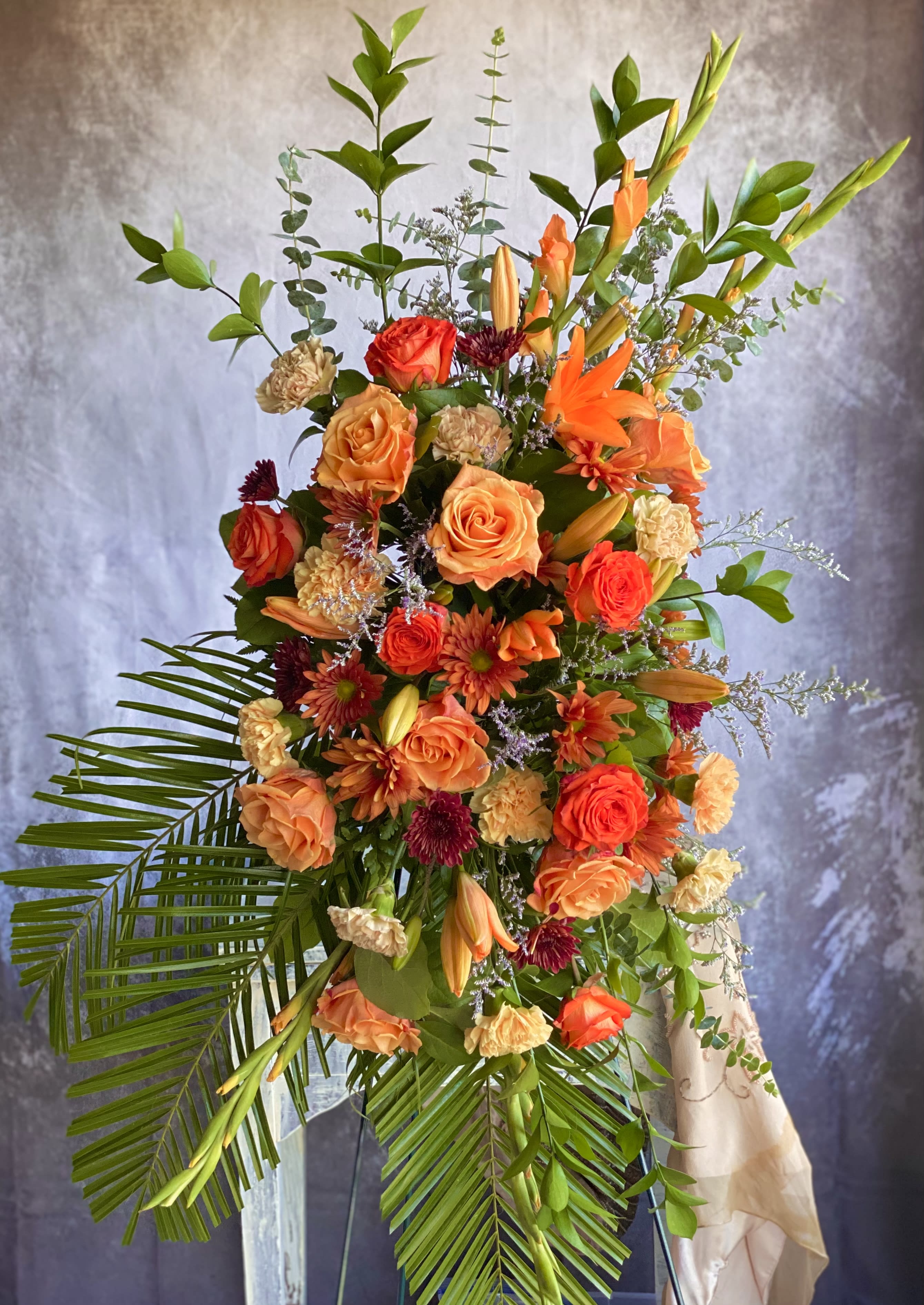 Tall orange and peach standing floral spray with roses and lilies on an easel.