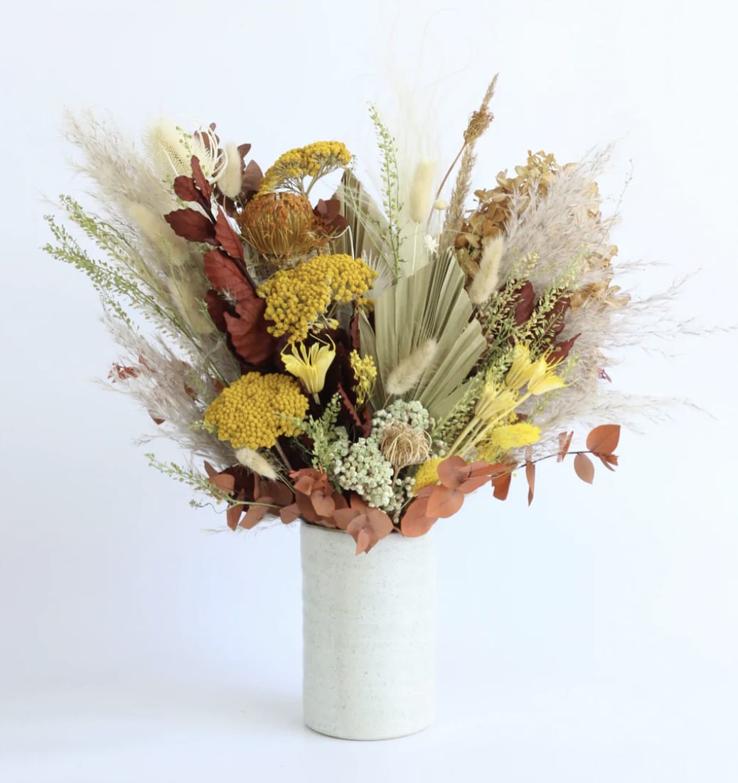 Tall dried-style arrangement with yellow and rust flowers in a white cylindrical vase