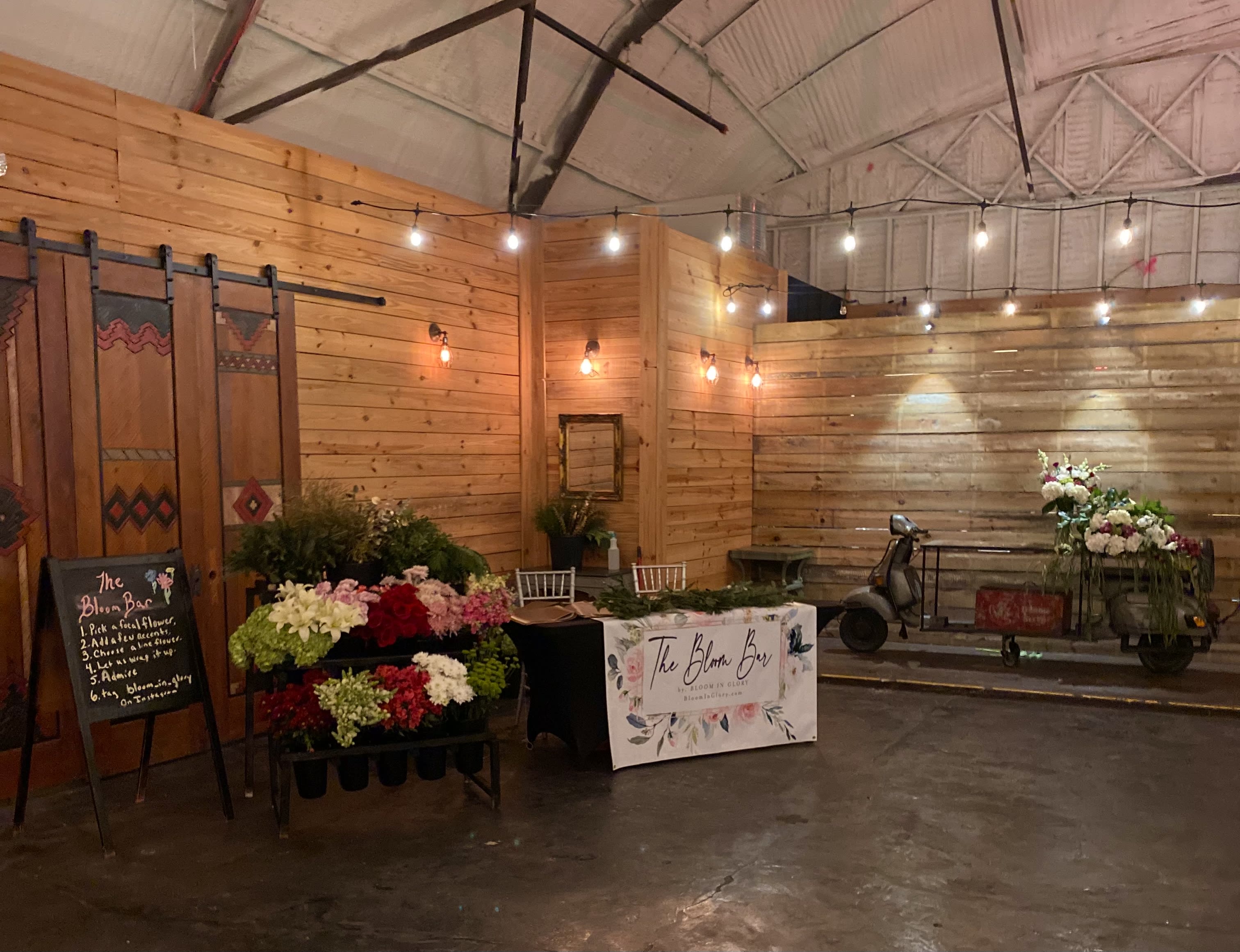 Rustic indoor flower bar setup with racks of mixed blooms and a scooter decorated with white and pink arrangements.