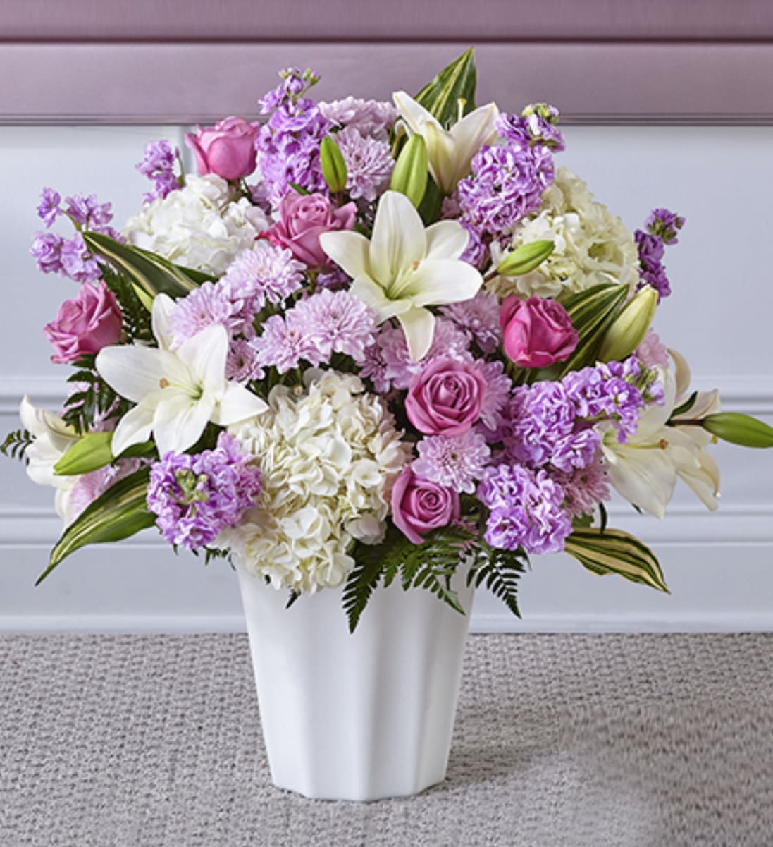 Tall arrangement of white lilies, pink roses, and lavender flowers in a white vase