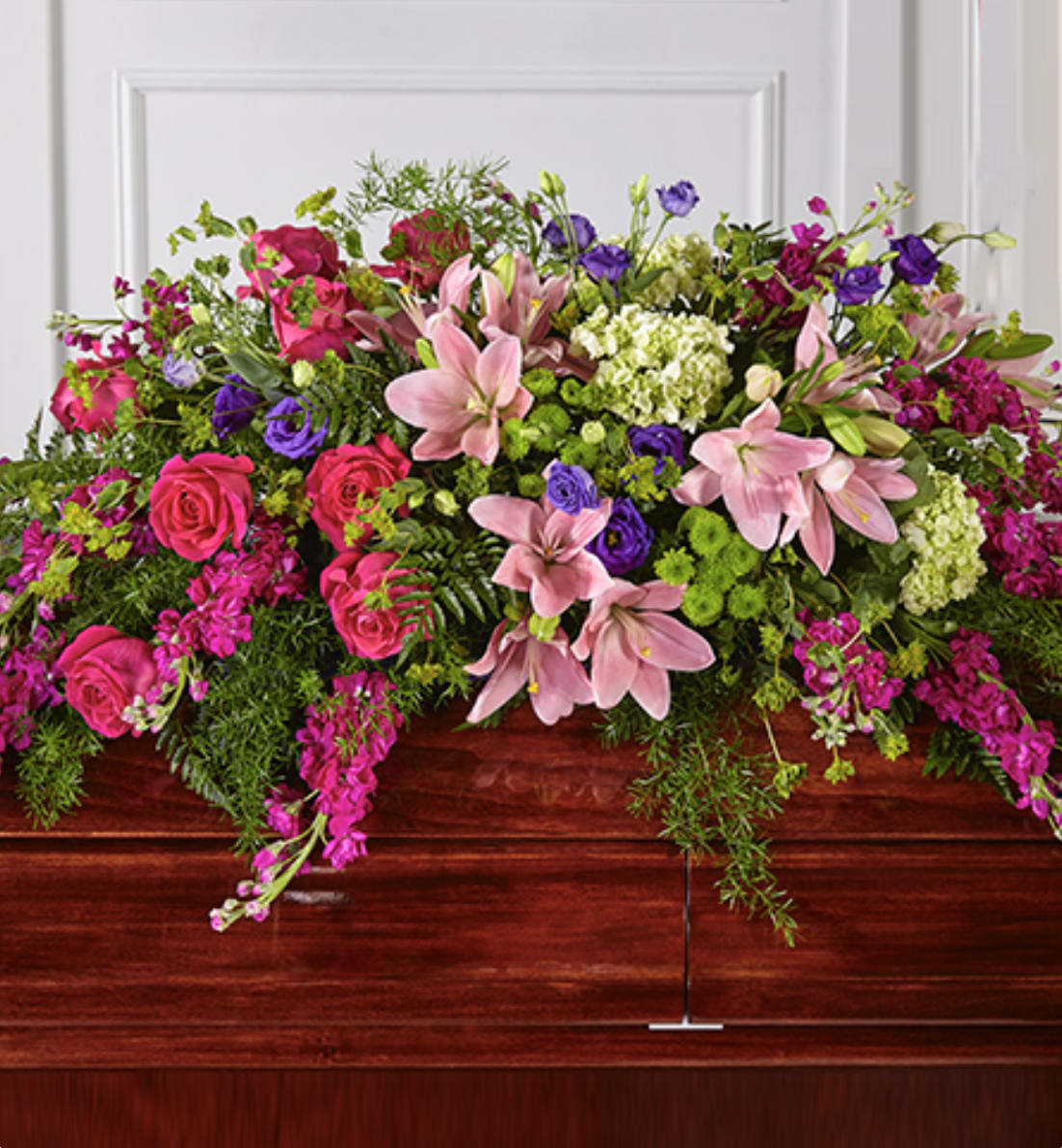 Bright pink lilies, roses, and purple blooms arranged as a casket spray on a wooden casket