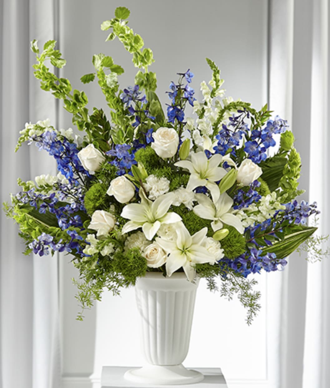 Tall white and blue arrangement with lilies, roses, delphinium and Bells of Ireland in a white pedestal vase.