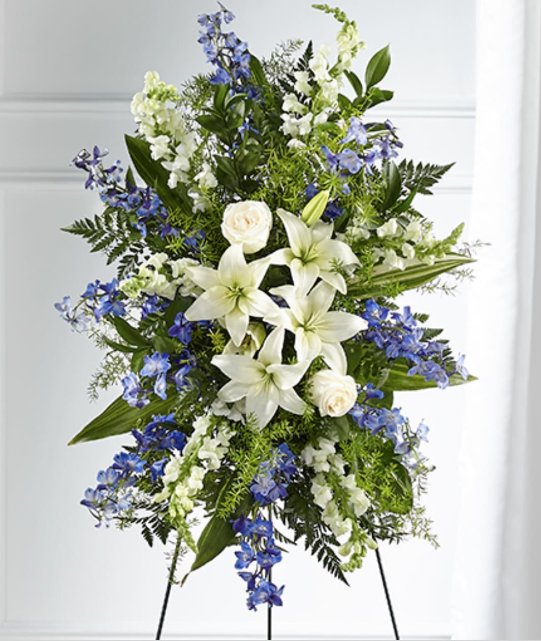 Tall standing spray of white lilies, roses, and blue flowers on a metal easel
