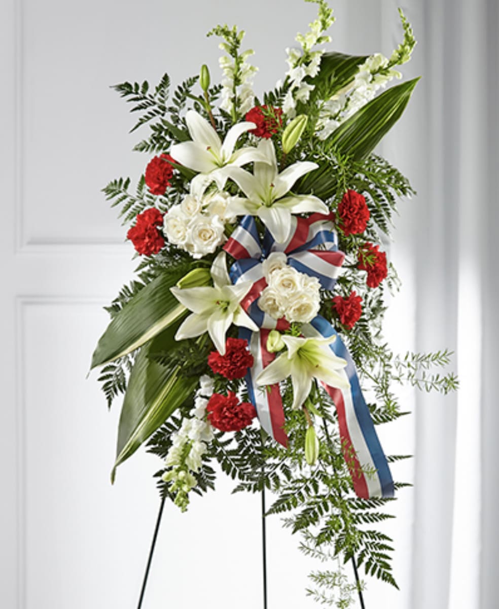 Tall standing spray with white lilies, white roses, red carnations and a red, white, and blue ribbon on an easel