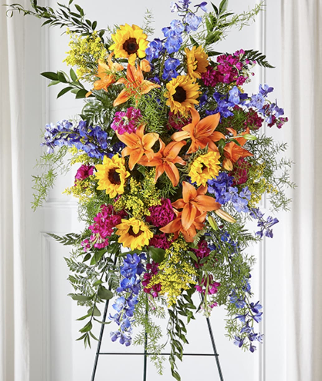 Tall standing spray with sunflowers, orange lilies, blue delphinium, and bright pink blooms on an easel