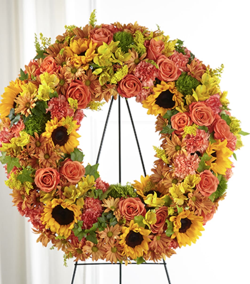 Standing floral wreath of yellow sunflowers, orange roses, and mixed warm-toned blooms on a metal easel