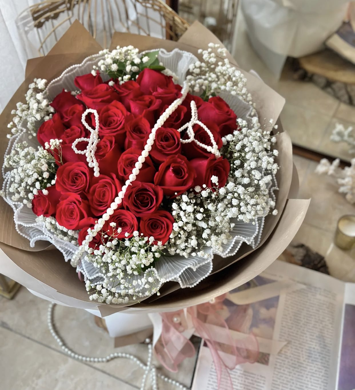 Hand-tied bouquet of red roses and white filler flowers wrapped in paper with draped pearl strands.