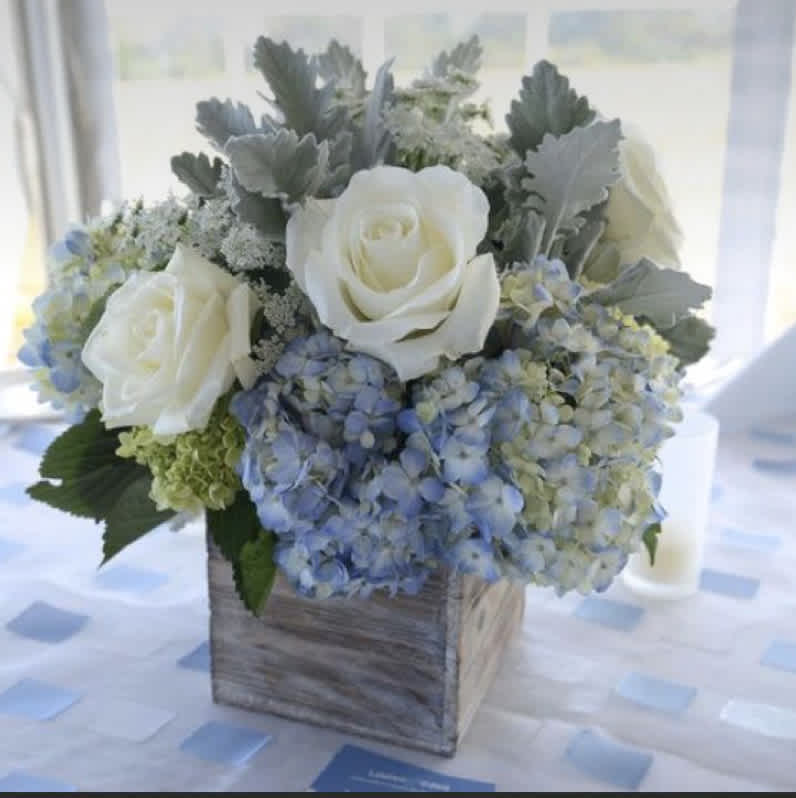 White roses and blue hydrangeas in a rustic wooden cube vase on a softly patterned table.