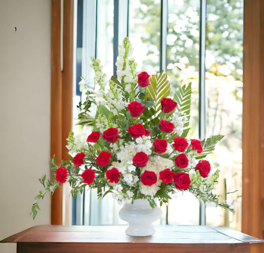 Tall arrangement of red roses and carnations with white flowers in a white urn on a table
