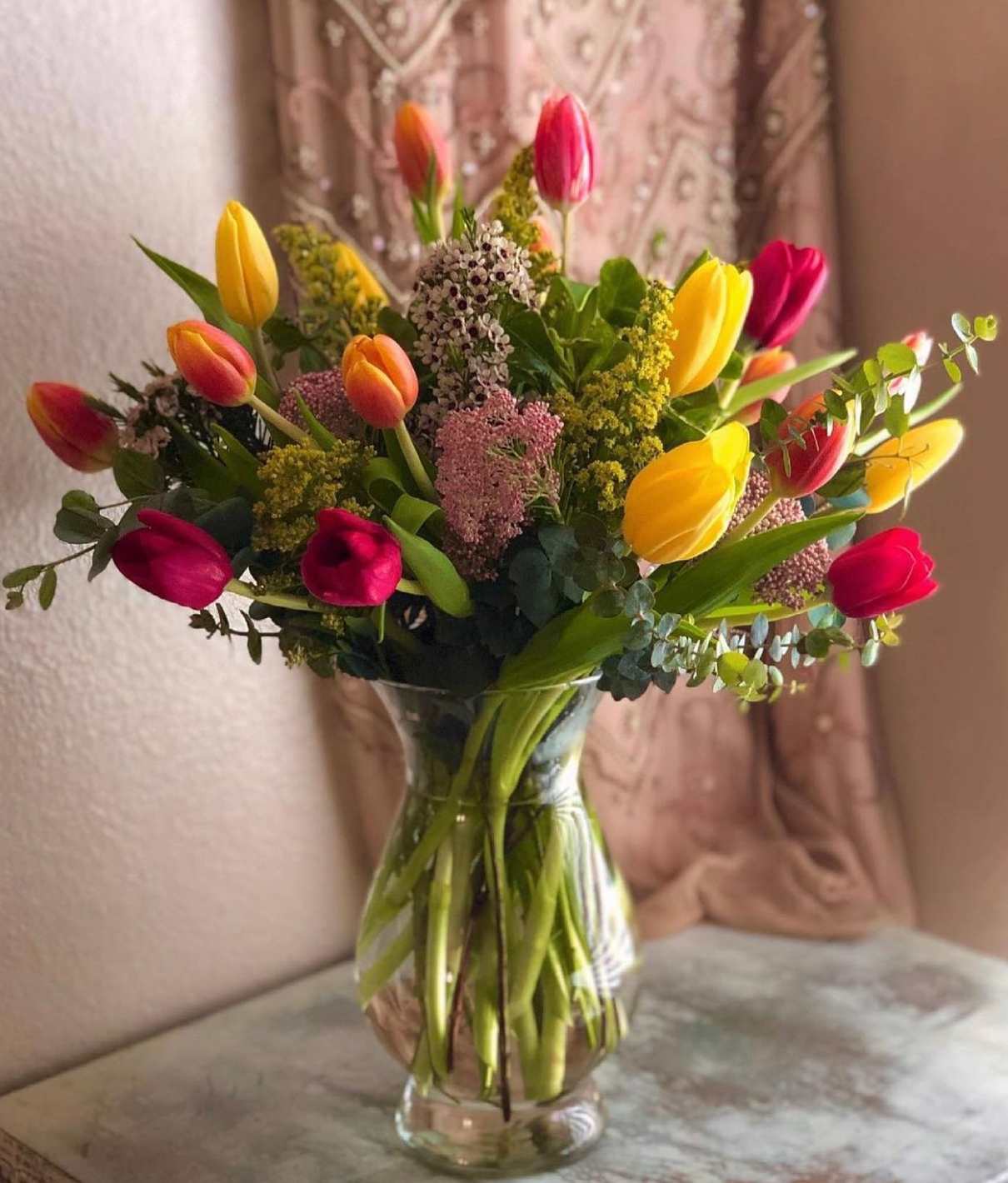 Clear glass vase of yellow, orange, and red tulips mixed with small filler flowers on a tabletop