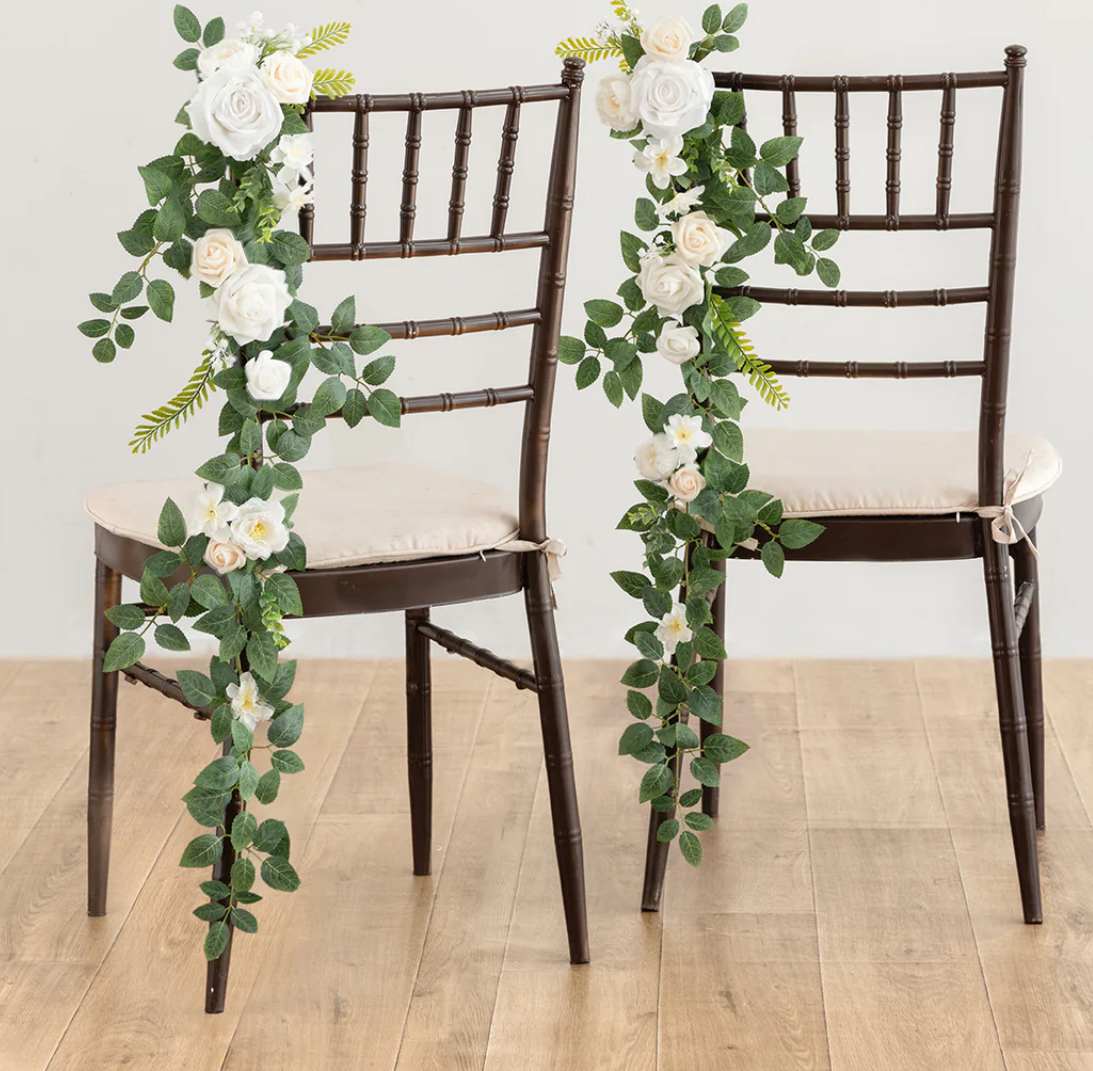 Pair of dark chairs decorated with cascading white and cream rose garlands with lush green foliage