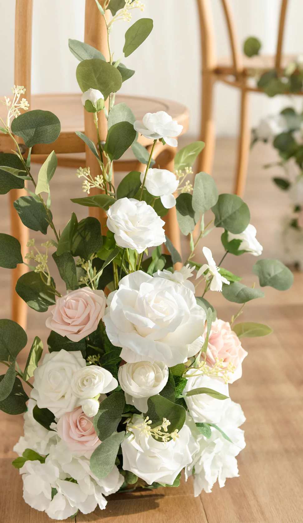 Floor arrangement of white and blush roses with abundant greenery beside wooden chairs