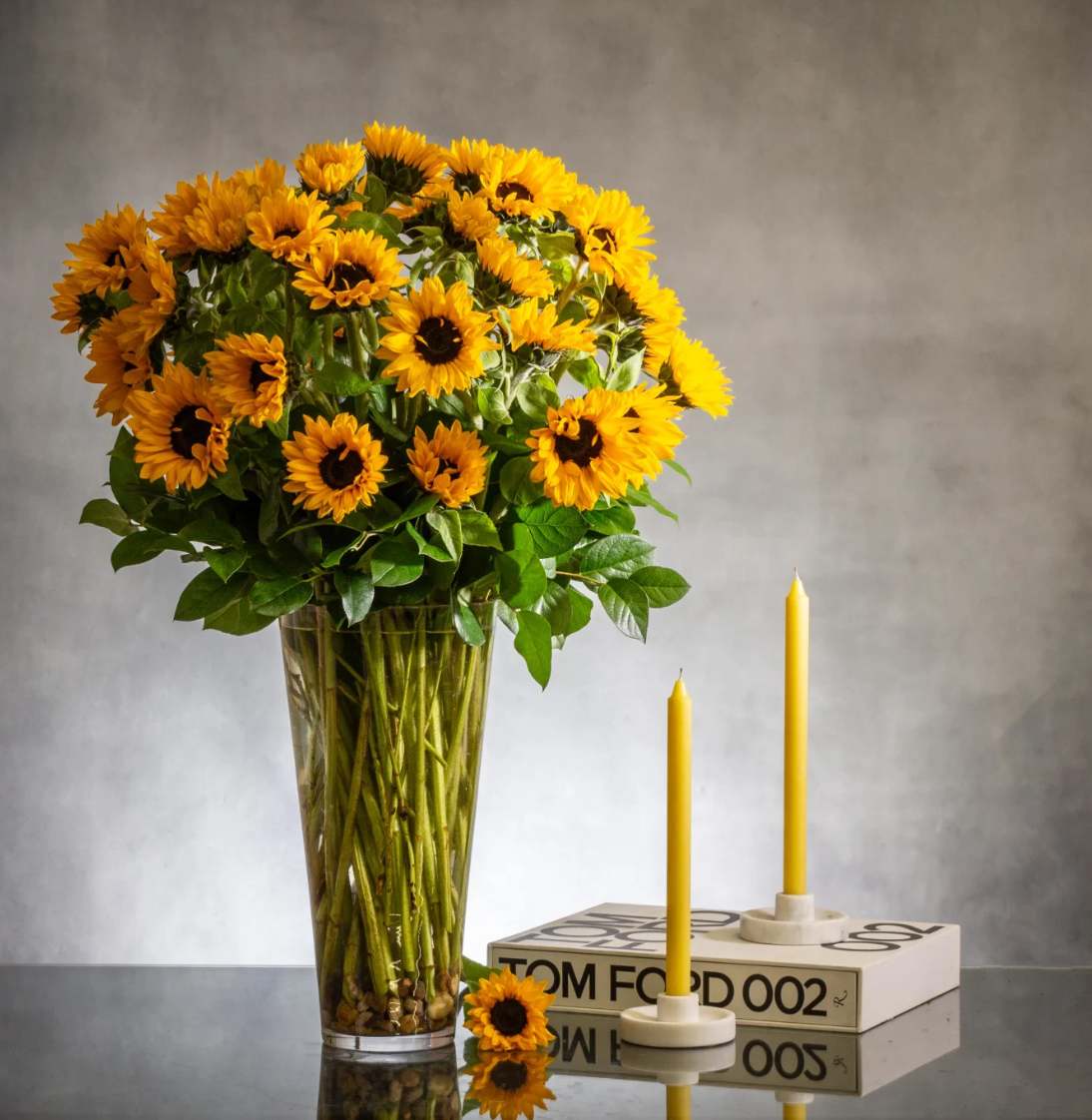 Tall arrangement of yellow sunflowers in a clear vase beside two yellow taper candles on a book.