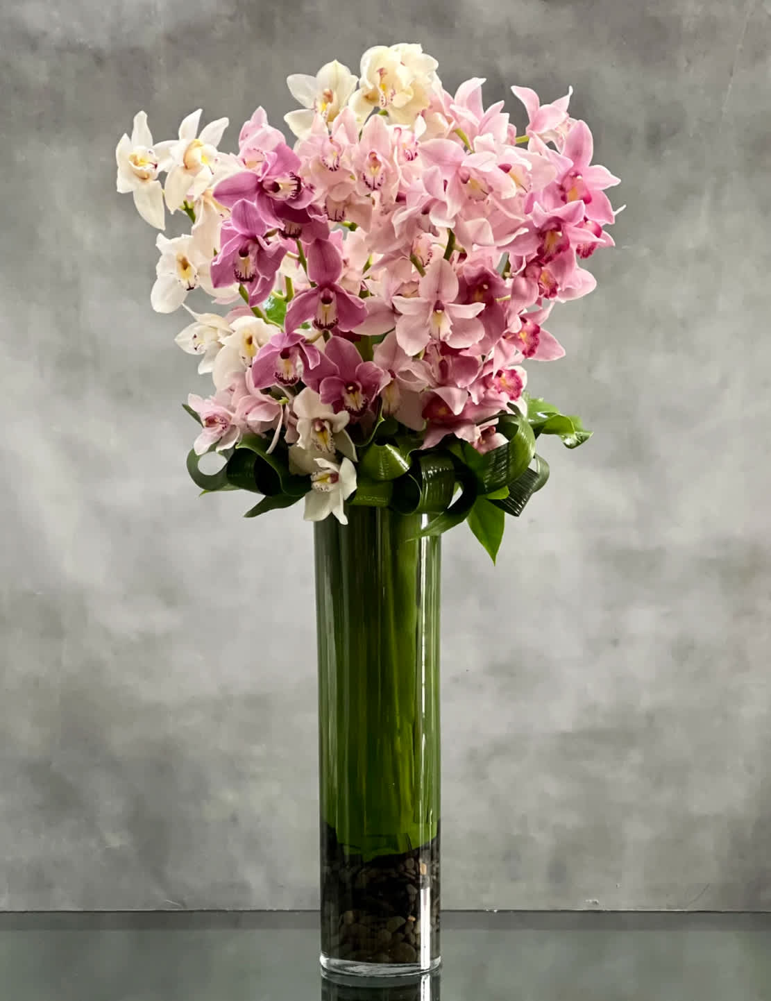 Tall cylinder vase of pink and white orchids with looped green foliage and stones at the base