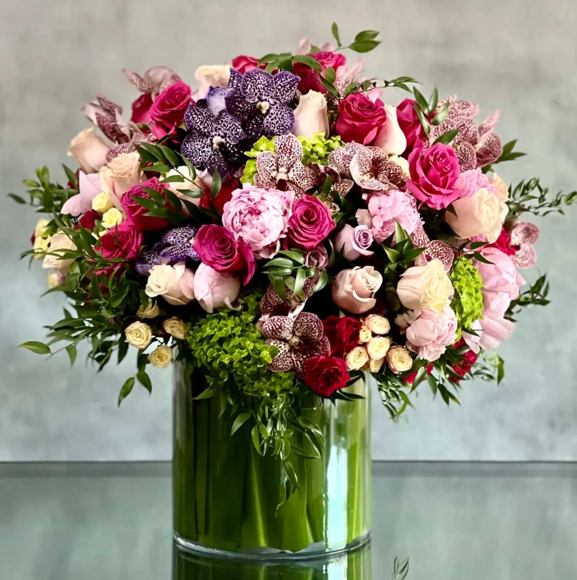 Large arrangement of pink roses, peonies, orchids, and green hydrangeas in a clear glass cylinder vase