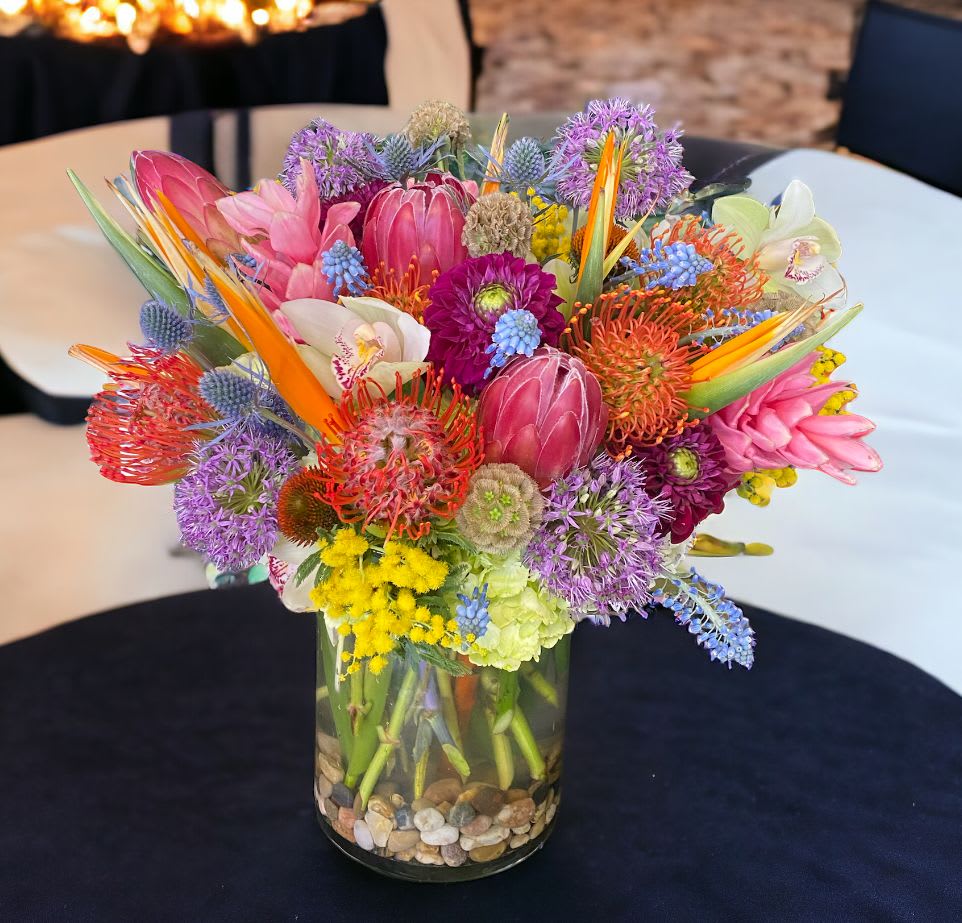 Bright mixed tropical and protea flower arrangement in a clear glass vase with stones.