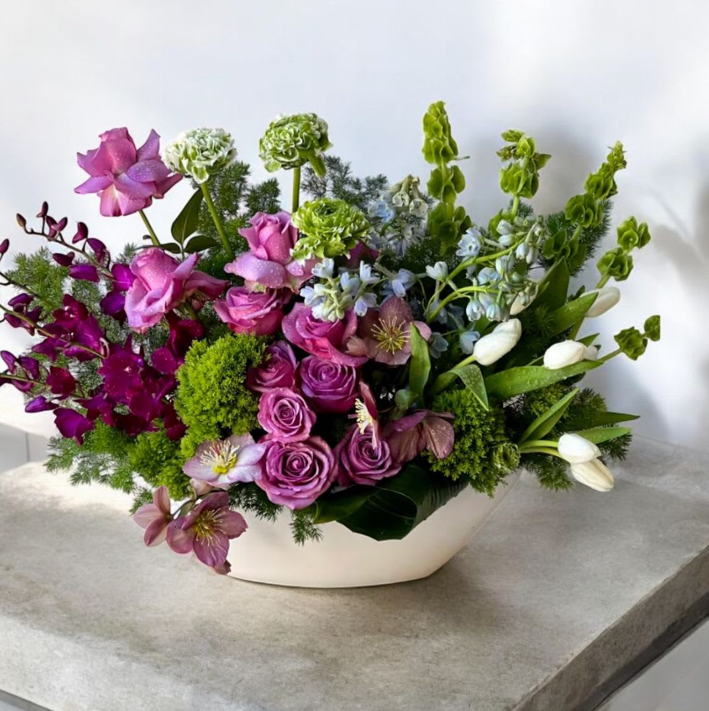 Low white ceramic arrangement with purple roses, orchids, and white tulips in an asymmetrical design