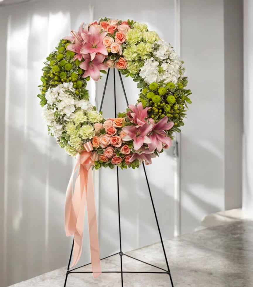 Floral wreath with pink lilies, peach roses, and white and green blooms on a black metal easel with peach ribbon